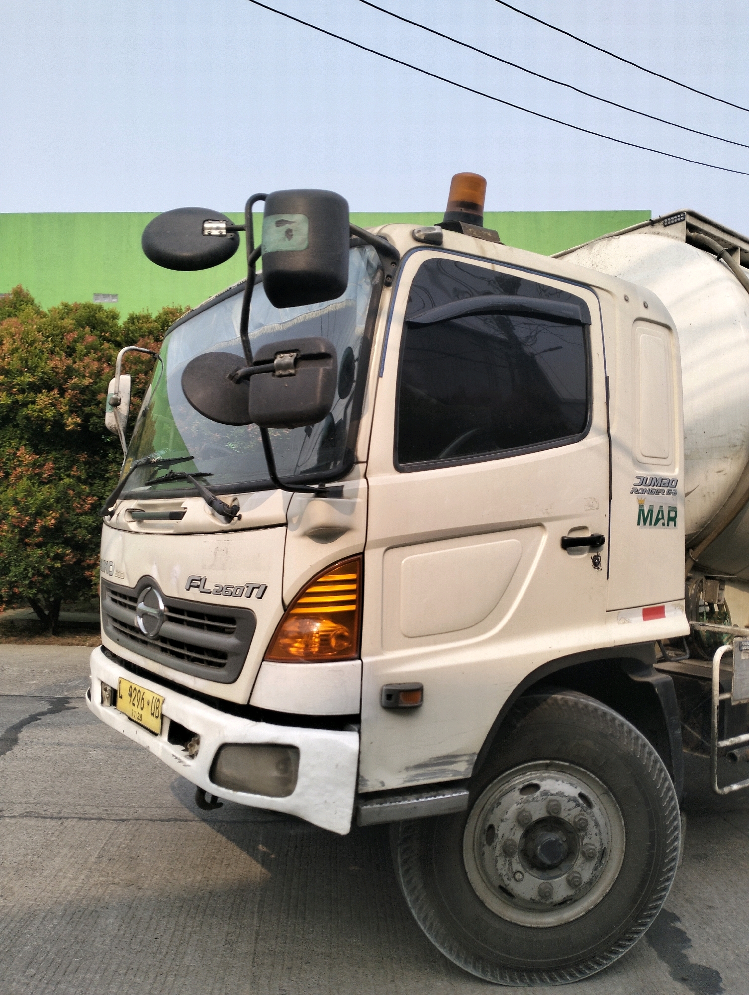 An outdoor, medium-shot, eye-level, frontal perspective captures a cream-colored cement mixer truck parked on a gray asphalt road. The truck is positioned diagonally, facing towards the left of the frame. The background features a bright green wall to the left, with a dense cluster of trees and foliage to the left and center. Power lines are visible in the upper portion of the sky, which is a pale blue.
The truck's features include a chrome grille with a circular HINO logo, amber turn signals on the front, and a license plate reading "L 9296 UB". The driver's side door has a black handle, and the windows are tinted. Above the windshield, a black plastic sun visor is attached. An orange beacon light is mounted on the roof of the cab. The side of the cab displays a logo with the words "JUMBO RANGER" and a crown graphic above the word "MAR". A large, black tire is prominent in the lower right quadrant of the image. There is a red and white reflective strip on the lower rear section of the truck's body.
The environment appears to be an urban or semi-urban area. The overall lighting suggests it is daytime, possibly late afternoon due to the angle of the shadows. There are no people visible in the image, and no visible interactions are occurring. The scene is static, with the truck stationary. The condition of the truck suggests it is operational, though some minor scuffs and dirt are visible on the bodywork. The location is specified as Gunung Sindur, Indonesia, which is a sub-district in Bogor Regency, West Java, Indonesia.