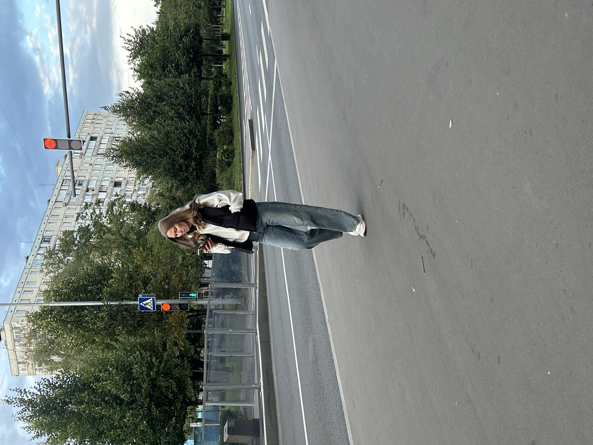 A young woman with blonde hair, wearing a white long-sleeved shirt, a black puffer vest, and light blue wide-leg jeans, walks across a pedestrian crossing on a cloudy day. She is looking down at her smartphone. In the background, there are trees, a building, and a traffic light showing red. The scene appears to be in Moscow, Russia, given the architecture and street markings consistent with Russian urban environments.