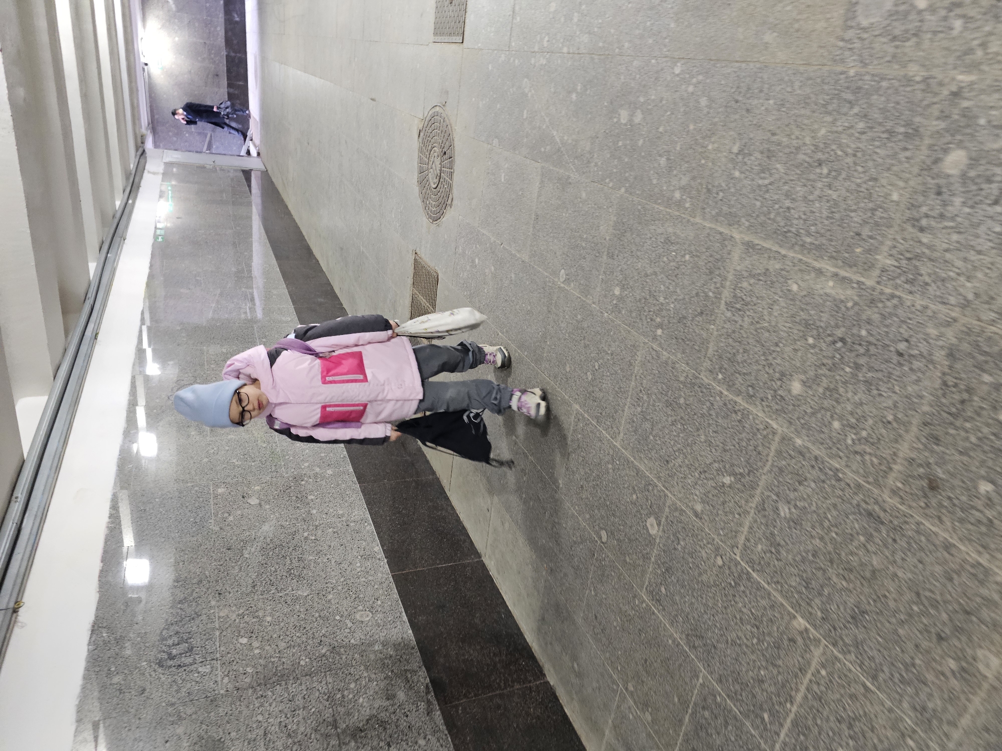 The image captures a young person standing in what appears to be an underground pedestrian walkway or subway station in Moscow, Russia. The person is wearing a light blue knit hat, eyeglasses, a pink and gray puffy jacket, gray pants, and sneakers. They seem to be holding a dark-colored bag. The floor is covered in gray square tiles, and there is a visible circular maintenance access cover. The lighting is somewhat dim, creating shadows. In the background, another person is seen walking further down the walkway. The walls are paneled, suggesting the modern design of the station. The overall scene suggests a typical moment of everyday life in a public transport area in a metropolitan city.