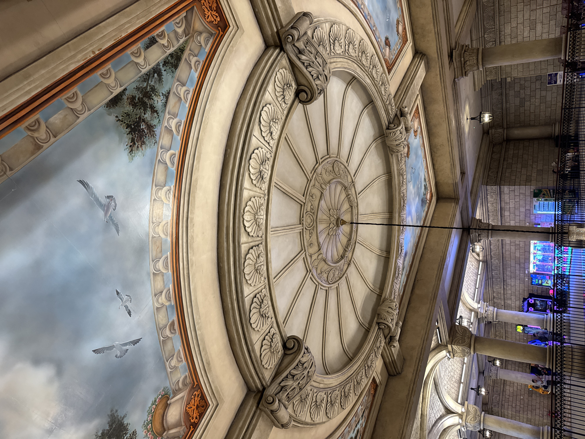 The image is a low-angle shot of an ornate ceiling in what appears to be an interior space in Đà Nẵng, Vietnam. The ceiling features a circular, decorative element with radiating lines and floral designs. Adjacent to the central circle, there is painted artwork depicting a sky with birds. The architecture includes columns with decorative capitals. To the right, there are visible walls made of stone blocks. A railing and electronic gaming machines suggest this may be a commercial or entertainment venue.