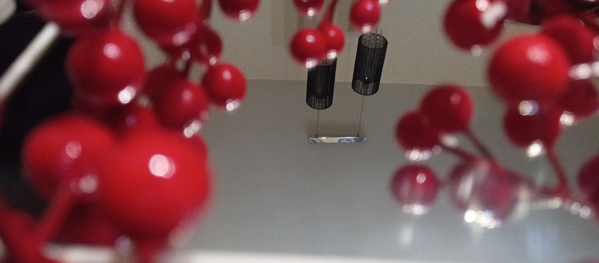 The image presents a close-up, shallow depth of field shot, creating a bokeh effect. The foreground is dominated by numerous vibrant red, spherical objects, possibly decorative berries or baubles, arranged in clusters on thin stems. These red elements are significantly out of focus, appearing as soft, glowing circles with bright white highlights, and frame the top, sides, and bottom of the composition.
In the background, two black cylindrical pendant light fixtures are visible, hanging from a light-colored, possibly grey or beige, ceiling or wall. These fixtures appear to have a textured or meshed exterior and are suspended by thin wires. They are connected to a reflective, horizontal silver-toned base. The background elements are also out of focus, though less so than the immediate foreground, suggesting a greater distance from the camera than the red objects.
The scene depicts an indoor setting, likely a room with modern lighting fixtures. There is no activity taking place, and no people or visible text are present. The lighting appears to be artificial, highlighting the reflective surfaces of the red spheres and the metal light fixture base.