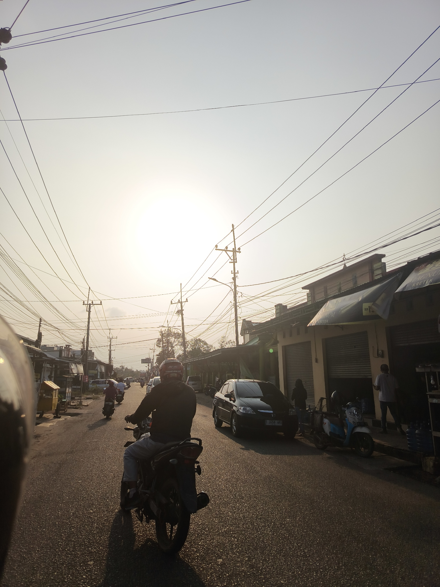 The image depicts a street scene in Pangkalpinang, Indonesia, during daylight with the sun positioned high in the sky, casting long shadows. The perspective is from a moving vehicle, likely a motorcycle, as evidenced by the partial view of a helmet in the foreground. The street is paved and appears to be a two-way road with several vehicles present. In the center of the frame, a person is riding a motorcycle. To the right, a black sedan is parked, and a lighter-colored scooter with a blue and white seat is also parked. Several buildings line the street on both sides, appearing to be shops with closed shutters. Overhead, a complex network of electrical wires is strung between numerous utility poles, creating a dense visual element in the sky. The sky is bright and hazy, suggesting either a sunny day or possibly pollution. The overall impression is of a typical urban street with moderate traffic and commercial activity.