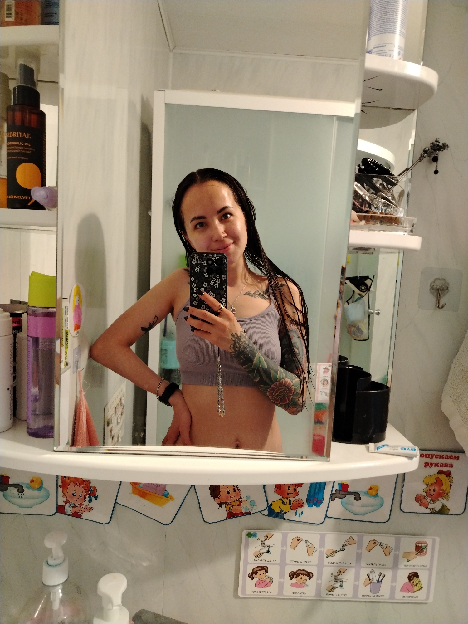 The image depicts a woman taking a selfie in a bathroom. She is fair-skinned with dark, wet hair. She is wearing a gray crop top and has numerous tattoos on her left arm and chest. She is holding a black smartphone with a floral design on the case.
The bathroom has a white cabinet and shelves stocked with various toiletries, including a bottle of "EBRIYAL" oil and other bottles with labels in Russian. The cabinet and shelves are decorated with images of cartoon children and instructions in Russian for washing hands and brushing teeth. There is also a towel hanging on a hook.
The location is likely in Noviy Urengoy, Russia, based on the Russian language text on the labels and decorations.