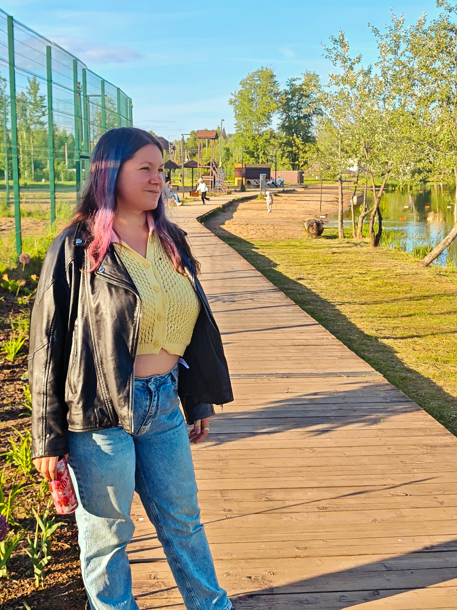 A woman with long, dark hair featuring pink and blue streaks stands on a wooden boardwalk in an outdoor recreational area in Zaprudnya, Russia. She is wearing a black leather jacket over a light yellow, knitted crop top, and light blue denim jeans. In her left hand, she holds a can that has "Mox" printed on its red and white design. She is looking towards the right side of the frame with a subtle smile, suggesting a pleasant disposition.
The setting appears to be a park or waterfront promenade. The boardwalk curves alongside a sandy/dirt path and a tranquil body of water, likely a lake or pond, reflecting the clear blue sky and surrounding trees. Lush green grass and various trees, some with light green foliage, line the water's edge and the path. In the background, distant figures can be seen on the sandy path, including one person in a white top walking, and others seated near rustic wooden structures that might be gazebos or playground elements. A tall green mesh fence is visible on the far left, suggesting a sports court or enclosed area.
The scene is illuminated by warm, golden sunlight, casting long shadows across the wooden planks, indicative of either late afternoon or early morning. The sky is bright blue with a few scattered white clouds, contributing to the bright and clear weather conditions.
