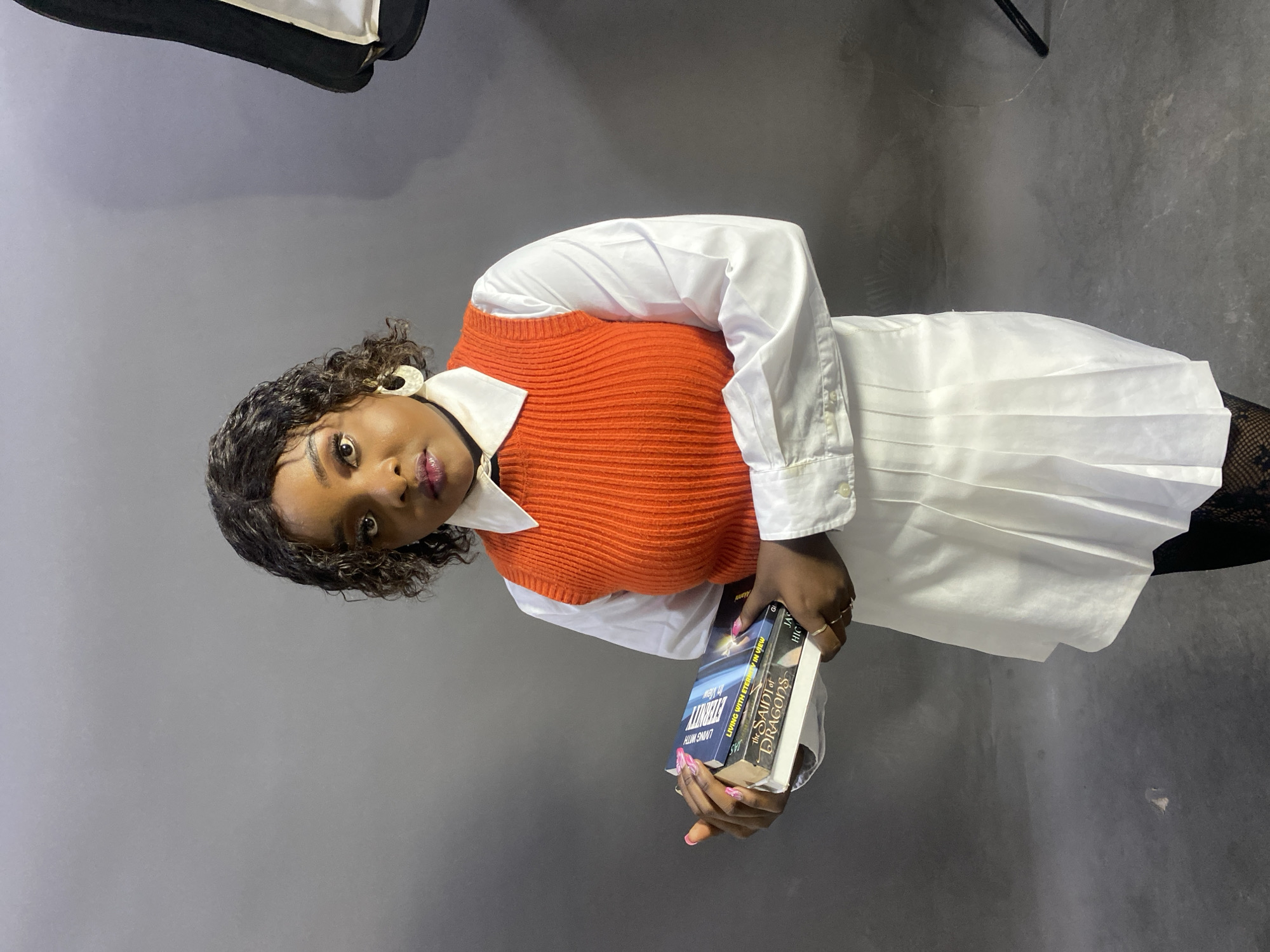 A young woman stands in front of a gray backdrop, posing with two books in her hands. She is wearing a white shirt dress under an orange knitted vest, and patterned tights, and accessorized with white earrings. The books she holds are titled "Living with Eternity in View" and "The Saint of Dragons". The location seems to be a studio or an indoor setting with lighting equipment visible in the upper left. There is no clear visual information to confirm the specific location in the city of Jos, Nigeria.