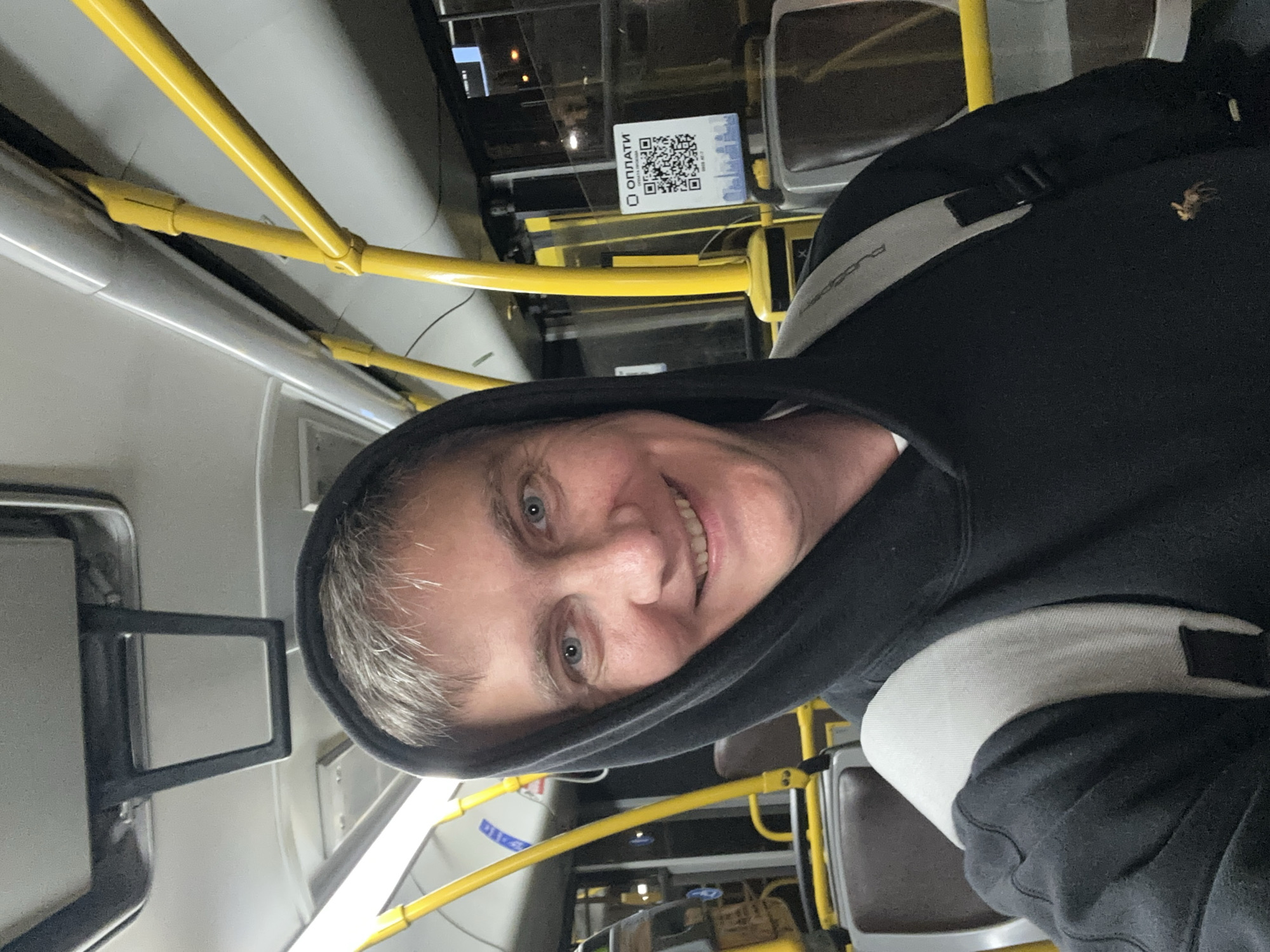 In this selfie taken from a low angle, a Caucasian man with short, graying hair and blue eyes smiles at the camera. He is wearing a black hooded sweatshirt with a grey backpack strap visible over his shoulder. The man is inside a public transportation vehicle, likely a bus or tram, given the presence of yellow handrails and brown seating. A QR code with Cyrillic text indicating "ОПЛАТИ" (which translates to "Pay") is visible on a poster behind him, suggesting a payment method for services within Minsk, Belarus. The background is blurred, but shows more handrails and seats, and the exterior lights of the city at night are visible through the windows.