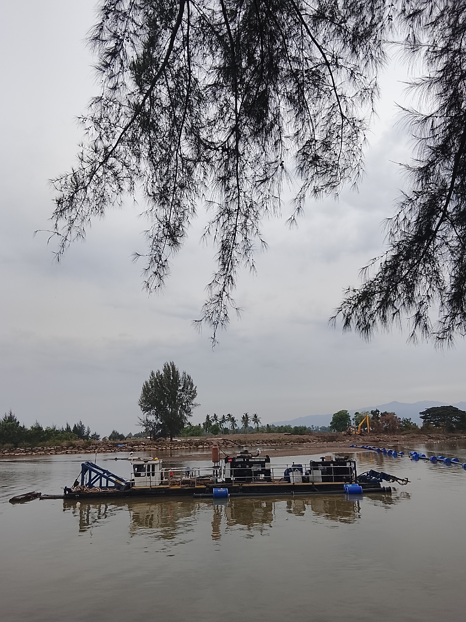 A dredging barge is anchored in the murky water of a river near Reudeuep, Indonesia. The barge, a long, flat-bottomed vessel, has a white cabin with windows at the front, and various machinery, including pumps and pipes, extend from its deck. A large blue pipe, supported by buoys, stretches from the barge towards the shore, suggesting ongoing dredging operations. In the background, the riverbank is lined with a rocky barrier, and beyond that, a dense cluster of trees and palm trees. Further inland, a yellow excavator is visible, indicating construction or land reclamation work. The sky is overcast, contributing to the subdued light of the scene. In the foreground, dark, needle-like branches of a tree overhang the water, partially obscuring the view of the sky and the far bank. The water's surface is calm, with subtle reflections of the barge and surrounding elements.
