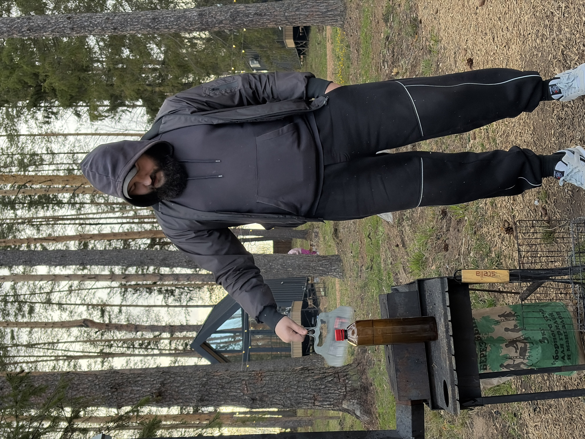 This outdoor image, taken in Мишкино, Russia, captures a man preparing for a barbecue or fire. The central figure, a bearded man wearing a dark hooded jacket, dark sweatpants with white stripes, and white sneakers, stands facing to his right, in the process of pouring liquid from a clear plastic bottle with a red cap into a brown glass bottle. The brown bottle is positioned on a metal barbecue grill stand, which features a charcoal grate.
The setting is a natural, wooded area, possibly a campsite or a property in a forest. Tall pine trees dominate the background, with their trunks visible against a light, overcast sky. The ground beneath is covered with dried leaves, pine needles, and patches of sparse grass. In the mid-ground, a modern-style cabin or house with large dark-framed windows can be seen amidst the trees, hinting at a leisure or residential context.
On the grill stand, a green bag of charcoal is visible, bearing Cyrillic text that reads "БЕРЕЗОВЫЙ" (Birch), "3 кг," and "ДЛЯ ГРИЛЯ И МАНГАЛОВ" (For grills and barbecues), along with silhouettes of people grilling. A wooden handle on the grill itself is inscribed with the word "stella" in lowercase. The man's white sneakers have a distinct black logo on the side. The activity suggests he is igniting or preparing to ignite a fire for cooking. The time of day appears to be daytime, possibly late afternoon, under diffused light conditions.