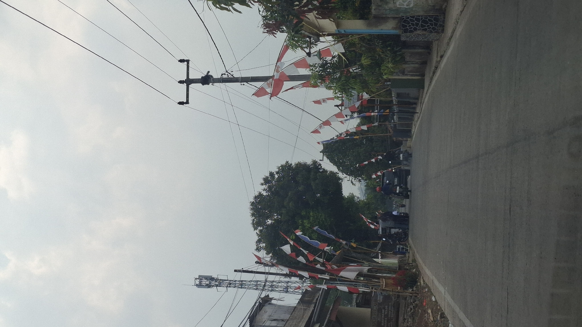 The image captures a street scene in Cibinong Hilir, Indonesia, characterized by a cloudy sky and an overcast atmosphere. Red and white Indonesian flags are prominently displayed along both sides of the asphalt road, adorning poles and extending over the street. On the left side of the road, a telecommunications tower stands tall, accompanied by a building with a corrugated metal roof. Several motorcycles are parked and in motion, with some individuals visible. A black car is also present on the road, with a driver and passenger. On the right side of the street, a utility pole with multiple wires runs overhead, and more flags are visible. Residential buildings with simple facades line the right side of the road, with some vegetation present. The road itself shows signs of wear, with some cracks and patches. The overall impression is a local community preparing for or celebrating a national event, with the flags serving as a clear indicator of Indonesian national pride.