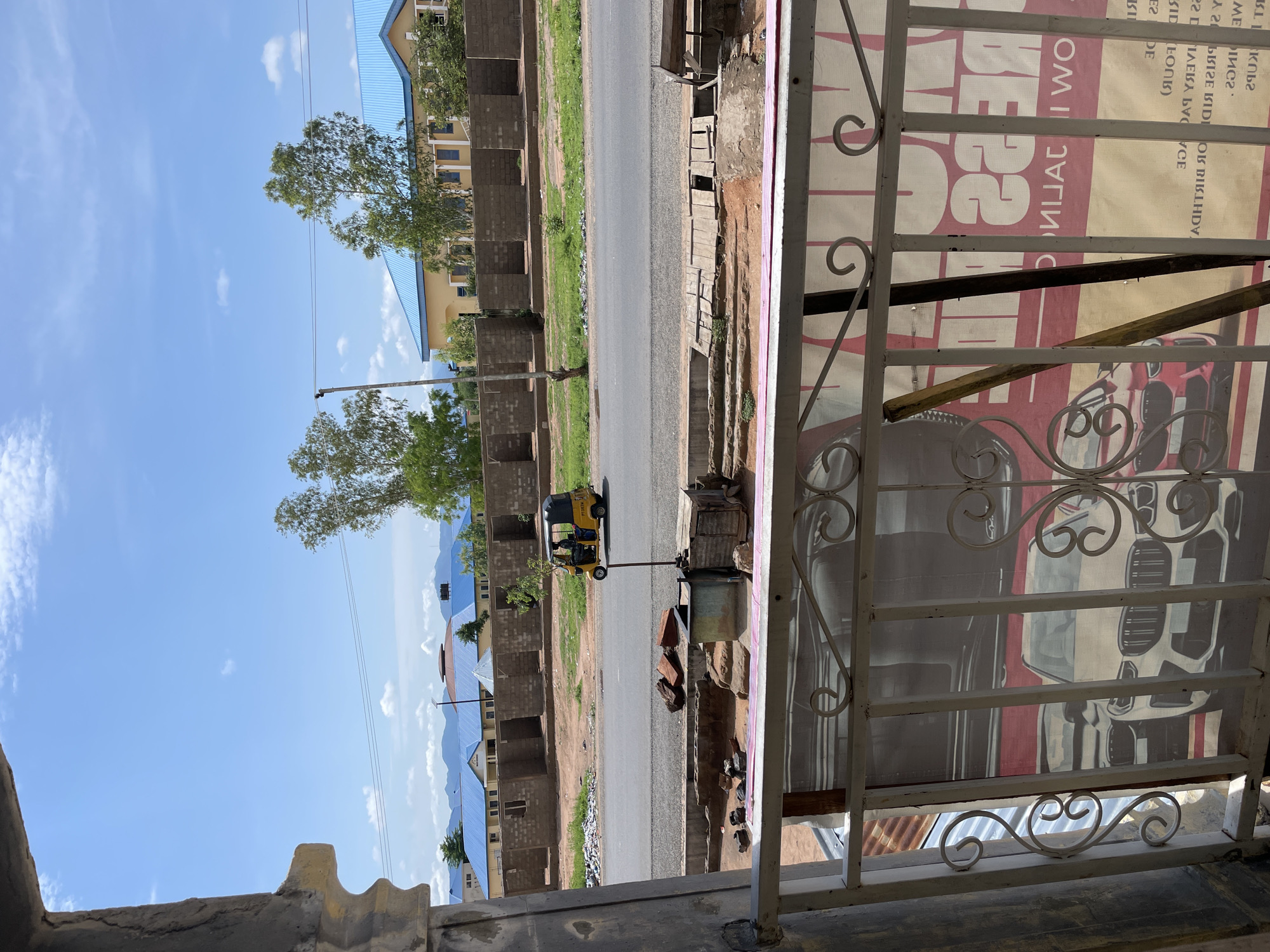 The image captures a detailed street-level scene in Jalingo, Nigeria, viewed from a slightly elevated perspective, possibly a balcony or window, framed by a white, ornate metal railing in the foreground.
The immediate foreground is dominated by a white, decorative metal railing featuring spiral and scroll motifs. Behind this railing, a large, rectangular advertising banner is partially visible. The banner displays the front of a white SUV, identifiable by its distinctive grille, and a smaller section of a red vehicle. Reversed text, including the numbers "22" and the word "PRESS," is discernable on the banner. To the left, beneath the railing, a section of a corrugated metal roof is visible.
In the midground, a two-lane paved road traverses horizontally across the frame. A yellow three-wheeled auto-rickshaw, commonly known as a Keke Napep, is depicted in motion on the road, traveling from right to left. The driver, a single individual, is visible within the vehicle. Adjacent to the road, on the near side, is an unpaved dirt strip containing scattered rubble, loose red bricks, and a makeshift wooden covering over a drainage channel. A slender wooden pole stands upright on this strip.
The background features a row of unfinished structures constructed from concrete blocks, each appearing as a separate unit with an open front facing the road. Beyond these, several larger, completed buildings are visible, distinguished by their bright blue corrugated metal roofs. One building in the distance exhibits a central, rounded, dome-like roof structure. Green-leafed trees are interspersed throughout the background, growing around and between the buildings. The far distance reveals a range of faint blue mountains or hills under a bright blue sky dotted with scattered white cumulus clouds. Utility lines are visible traversing the upper portion of the sky. The overall scene depicts a sunny day in an active, developing urban or semi-urban area.