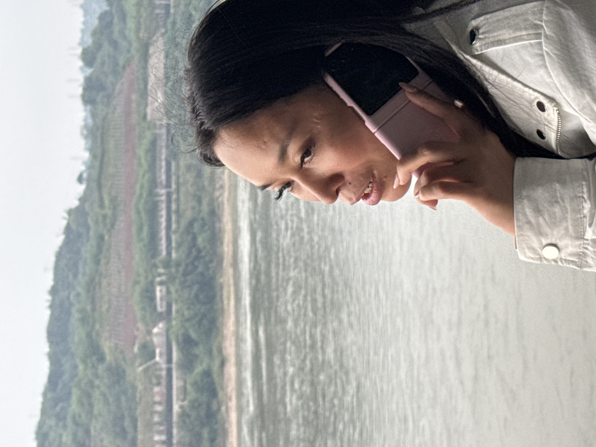 A woman is holding a pink smartphone to her ear. She is wearing a light-colored denim jacket. Behind her is a view of a river and a tree-covered hillside. The location is the city of Jiangjin, China.