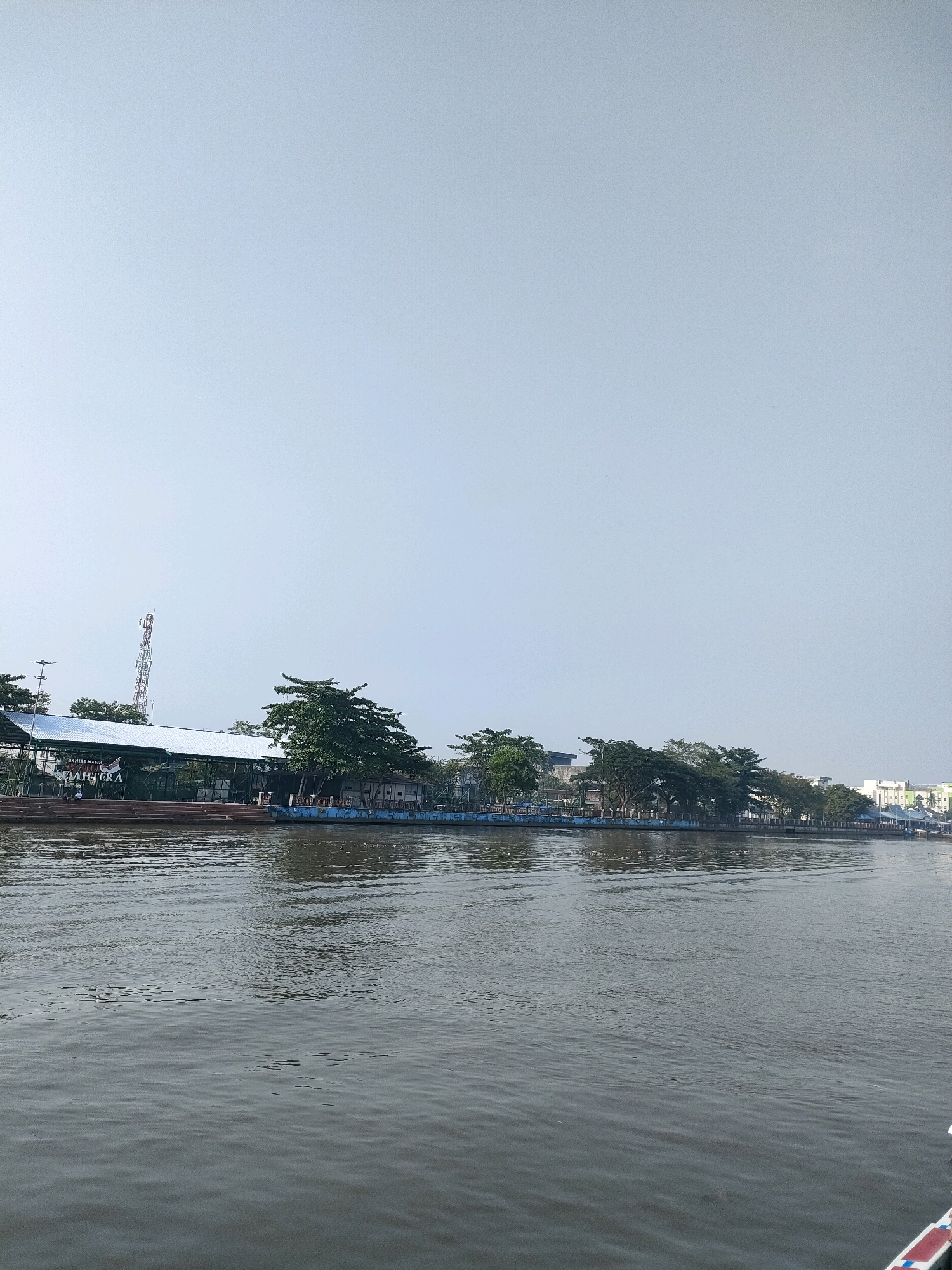 The image shows a wide, calm river flowing through an urban area, likely Banjarmasin, Indonesia. Along the riverbank, there is a row of trees and a building with a large, white roof. The building appears to be a restaurant or a public facility, with tables and chairs visible outside. A red and white antenna tower stands tall in the background, suggesting proximity to communication infrastructure. The sky is a pale blue, indicating a clear day. The water reflects the surrounding environment, creating a serene atmosphere. In the foreground, a small portion of a boat is visible, suggesting the image was taken from a boat on the river. The overall scene depicts a peaceful riverside setting within a city, characteristic of areas in Banjarmasin known for their riverfront developments and recreational spaces.