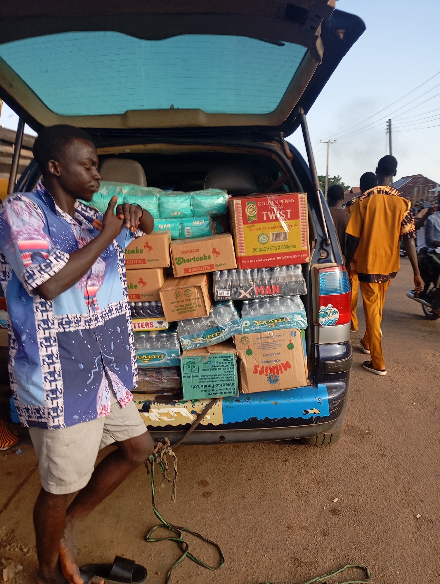 A full shot captures a man in a blue and white patterned shirt and light-colored shorts standing next to the open rear of a dark-colored vehicle, filled with various items. The man is looking to his left, with his hands raised and fingers spread, as if gesturing. Behind him, stacked inside the vehicle, are packages of diapers, boxes of biscuits, and bottles of beverages. A box with "GOLDEN PENNY PASTA TWIST" is prominently displayed. Two other individuals, one wearing an orange outfit, are visible in the background, walking away from the vehicle. The scene appears to be outdoors, with a paved road and a few buildings in the distance, suggesting an urban or semi-urban setting, likely in Shendam, Nigeria. The lighting indicates it is daytime.