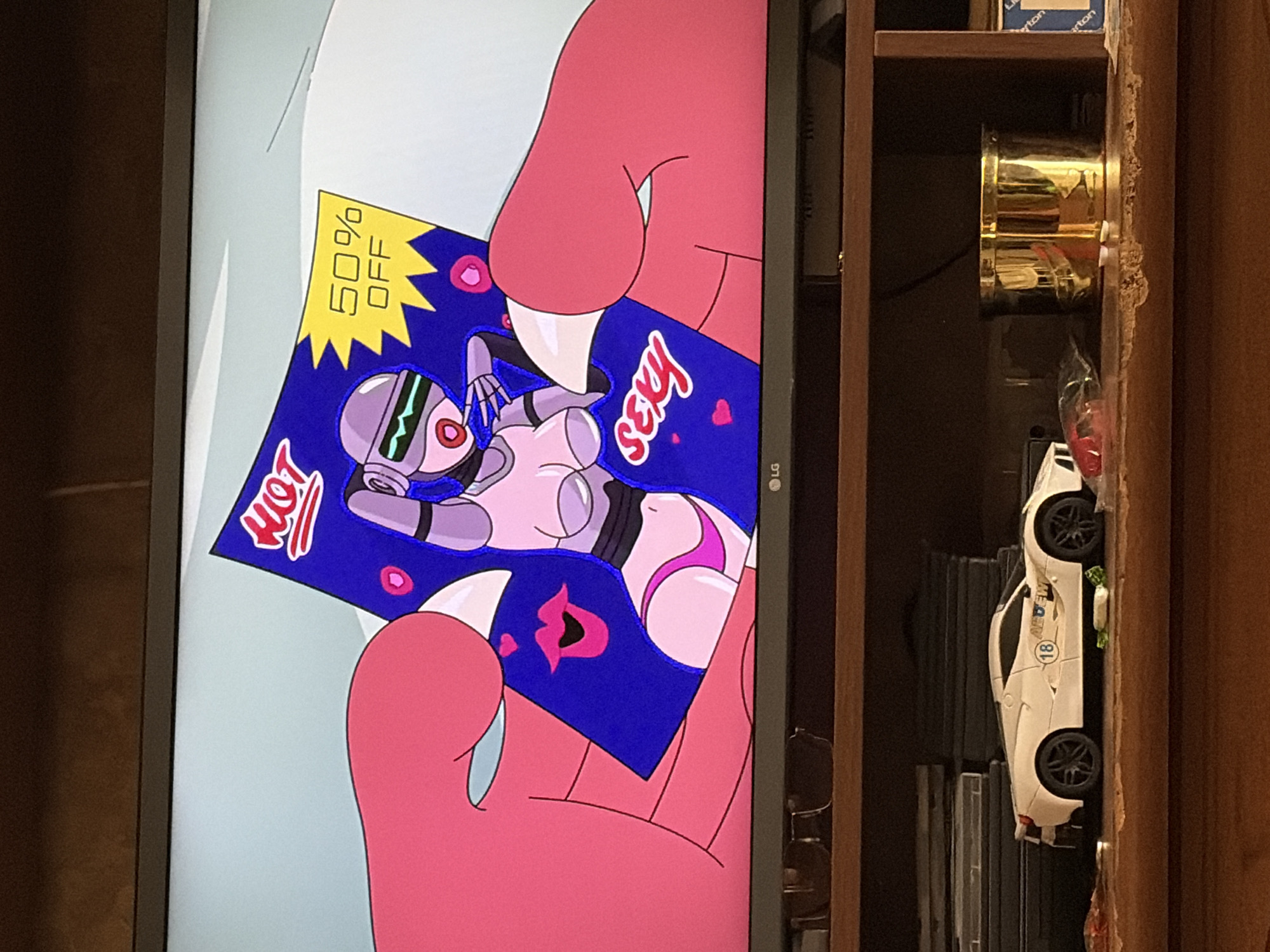An image shows a television screen displaying a cartoon advertisement for a "HOT SEXY" robot with "50% OFF". The advertisement features a pink-skinned character with large hands holding the card. Below the television, a shelf displays several items, including a white toy car and a golden cylindrical container. The scene appears to be an indoor domestic setting, possibly a living room. The background of the television screen is a light blue.