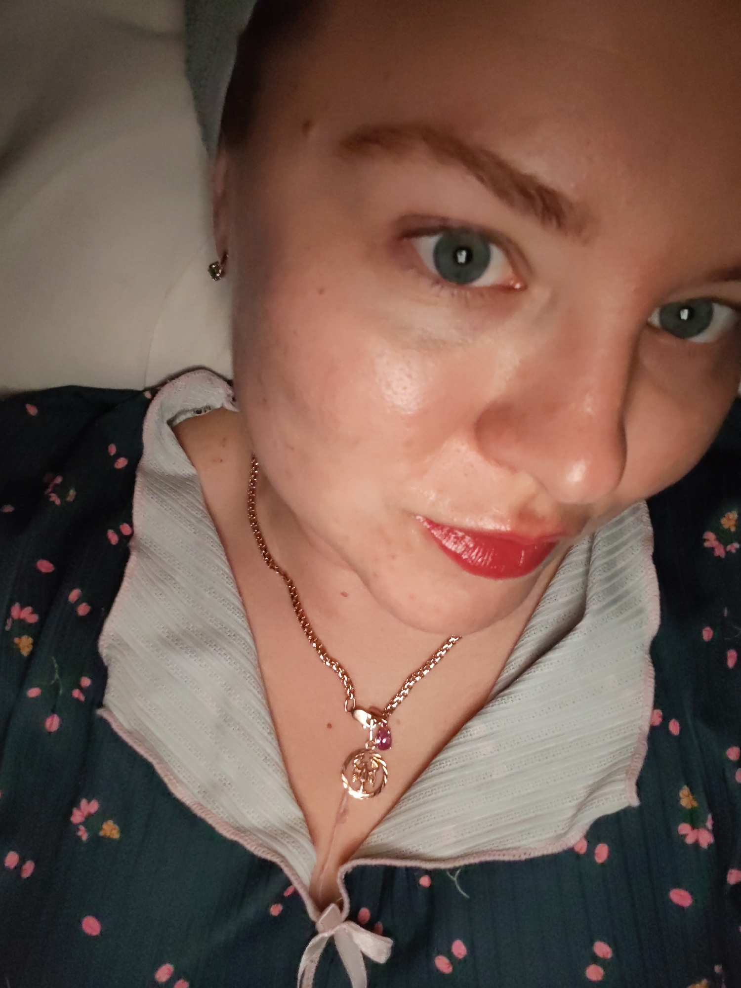 A person is seen in a close-up shot. They have fair skin, light eyebrows, and blue eyes. The person is wearing red lipstick. They are wearing a green floral patterned top with a white ruffled collar. A gold necklace with a charm is visible. The background is blurred. There is no location information present in the image.