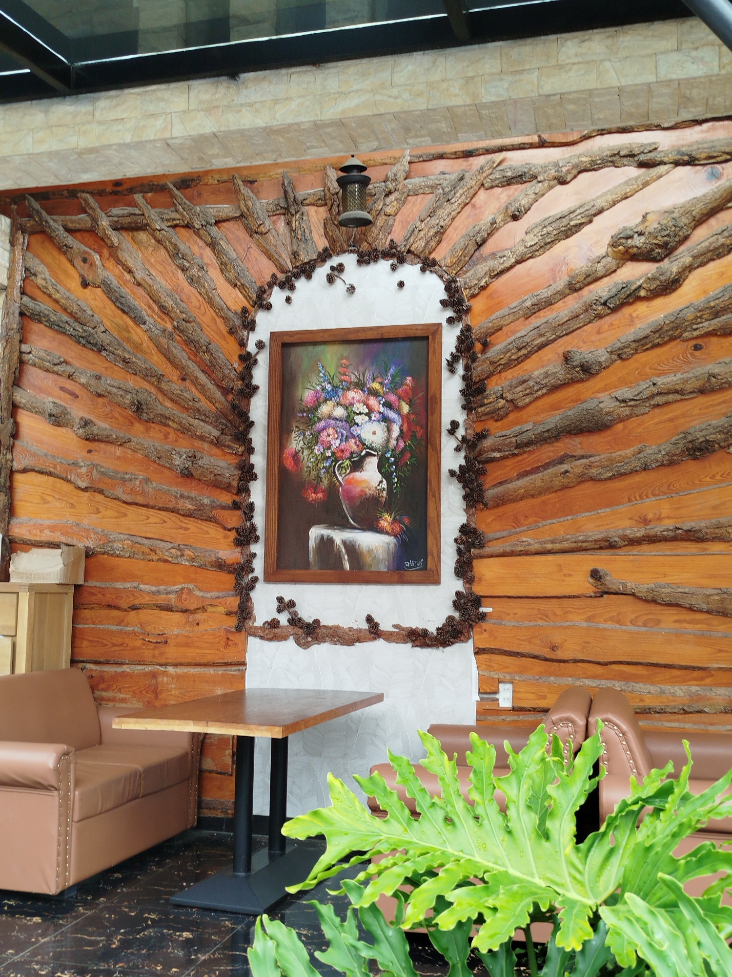 The image depicts an indoor seating area, likely a cafe or restaurant, with a rustic and naturalistic decor. The focal point is a large, ornate wall treatment made of horizontal wooden planks with rough-hewn branches strategically placed to form a sunburst or fan pattern radiating from a central arched alcove. Inside this alcove, a framed painting of a colorful bouquet of flowers in a vase is displayed. The alcove itself is outlined with small pinecones and twigs, adding to the natural theme.
The overall color palette is dominated by warm, earthy tones of wood and the vibrant colors of the painting. The lighting appears to be a mix of natural light from above and artificial light from a lantern mounted on the wall.
In the foreground, a lush green plant with large, deeply lobed leaves partially obscures a brown leather sofa. To the right of the sofa, a dark-legged wooden table with a simple rectangular top is positioned. Behind the table, another section of a brown leather sofa is visible. The flooring is a dark, glossy material with a marbled pattern.
The image also shows a glimpse of a stone-tiled wall and a reflective ceiling or overhead structure above the wooden wall. The style of decor, particularly the use of natural wood elements and the overall ambiance, is consistent with establishments in Da Lat, Vietnam, a city known for its pine forests, French colonial architecture, and a popular tourist destination that often features cafes with a rustic aesthetic.