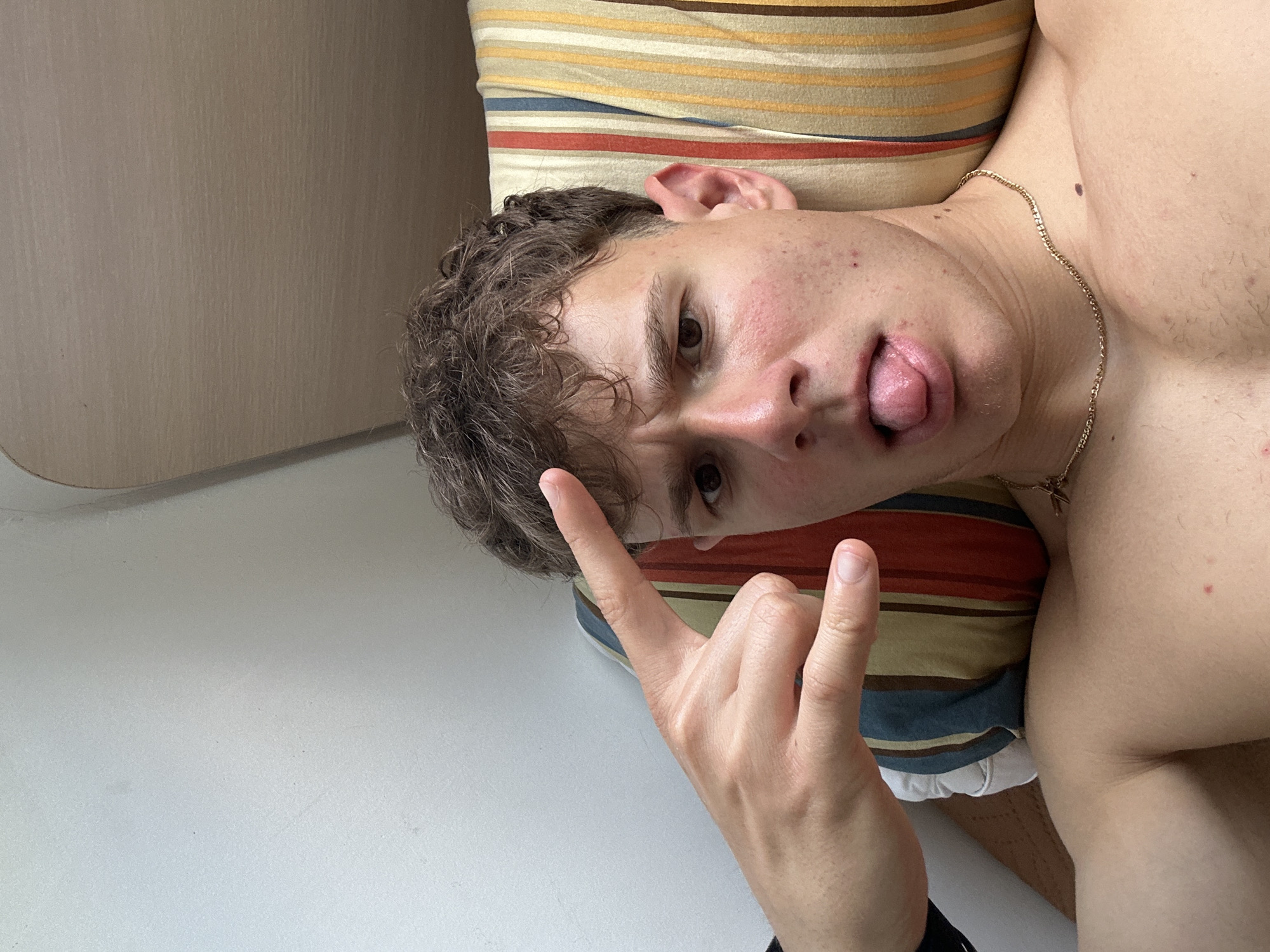 The image depicts a young, light-skinned individual with curly brown hair lying on a pillow with multi-colored horizontal stripes. The person is making a "rock on" hand gesture. The person is also sticking their tongue out. A gold chain is visible around their neck. The background includes a white surface and a light-colored wall. The location is unknown.