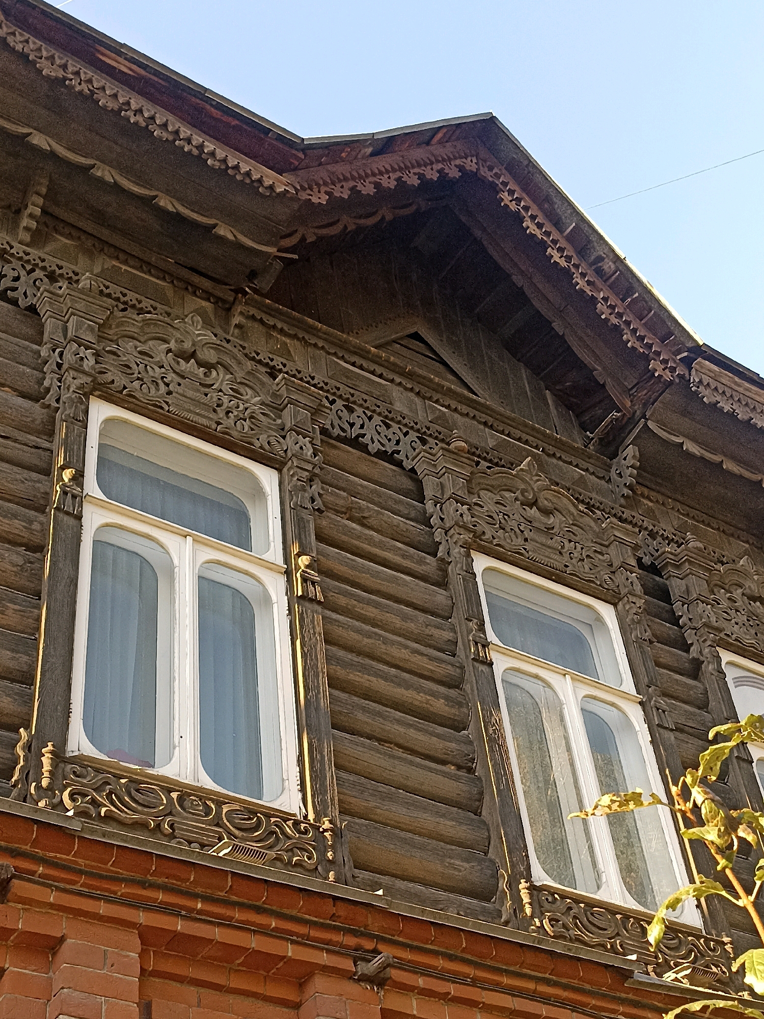This is a low-angle, full shot of a weathered, dark brown wooden building with ornate, carved wooden trim and arched windows in Novosibirsk, Russia. The building's log construction is visible, with horizontal logs forming the walls. The eaves of the roof are decorated with intricate, dark brown wooden carvings featuring floral and geometric patterns. Two white-framed, arched windows are prominent. The left window is larger and has two arched panes, while the right window, partially obscured by a leafy branch, also has an arched shape. The lower portion of the building visible in the frame is made of red brick. The sky above is a clear, pale blue. The image appears to be taken on a sunny day, as evidenced by the shadows and highlights on the wood and brick.