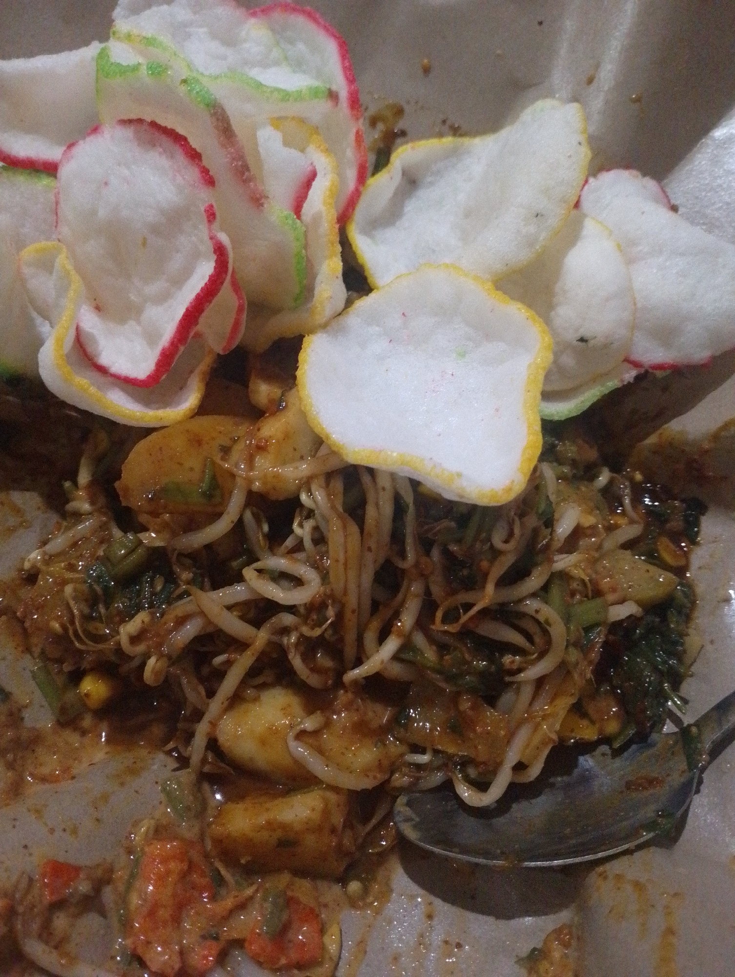 A close-up shot captures a serving of gado-gado, a popular Indonesian salad, presented on a bed of parchment paper. The dish features an assortment of blanched vegetables, including bean sprouts, and is generously coated in a thick, savory peanut sauce. Scattered atop the salad are several colorful krupuk, or Indonesian crackers, in shades of white, red, green, and yellow. A metal spoon rests in the lower right quadrant of the frame, partially submerged in the sauce. The background is dark and nondescript, focusing attention on the vibrant dish. This image was likely taken at a local restaurant or street food stall in South Tangerang, Indonesia, where gado-gado is a common and beloved culinary offering.