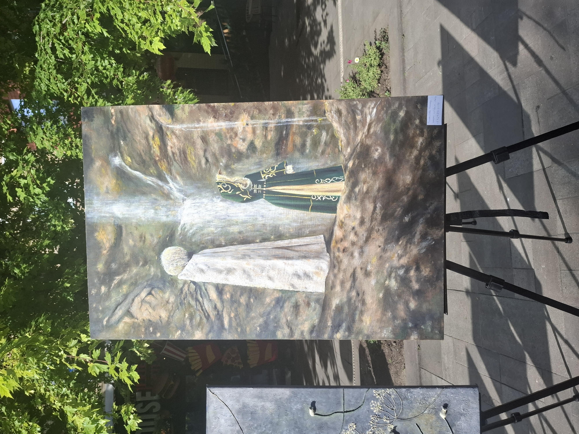 The image depicts an outdoor art exhibition in the city of Grozny, Russia. A large canvas painting, displayed on a black tripod easel, features a waterfall and two figures. One figure, in a white robe with a fluffy head, stands facing away from the viewer. The second figure, to the right, wears a green and gold traditional garment with intricate patterns. The painting's background suggests a rocky, natural setting. To the left of this painting, a portion of another artwork, a dark gray rectangular canvas with a delicate floral design, is visible, also on an easel. The exhibition is set against a backdrop of lush green trees and a paved walkway, with shadows cast by the trees and possibly buildings. In the background, obscured by foliage, there are indications of commercial buildings. A small plant with pink and white flowers is visible in the lower right foreground. A white label with handwritten text and a phone number is attached to the easel of the larger painting.