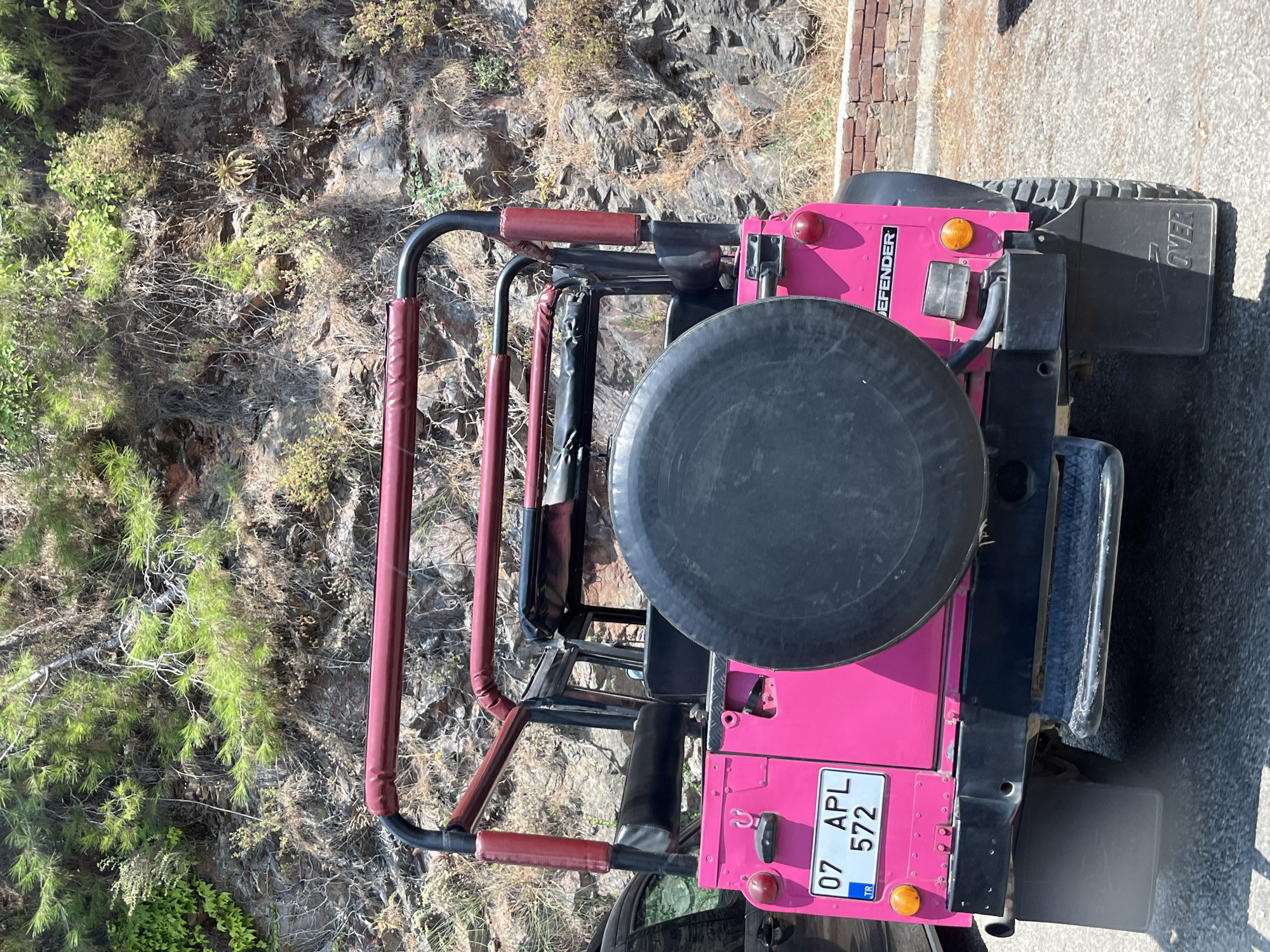 A bright pink Land Rover Defender with a spare tire mounted on the rear is parked on a paved surface. The vehicle has a black roll cage with red padding. A license plate reading "07 APL 572" is visible on the rear. The background consists of a rocky hillside with sparse vegetation and a low brick wall. The scene appears to be in a natural or semi-natural environment, potentially on the outskirts of a city. The sunlight suggests it is daytime. The image does not contain any people or discernable actions. The location is identified as Alanya, Turkey.