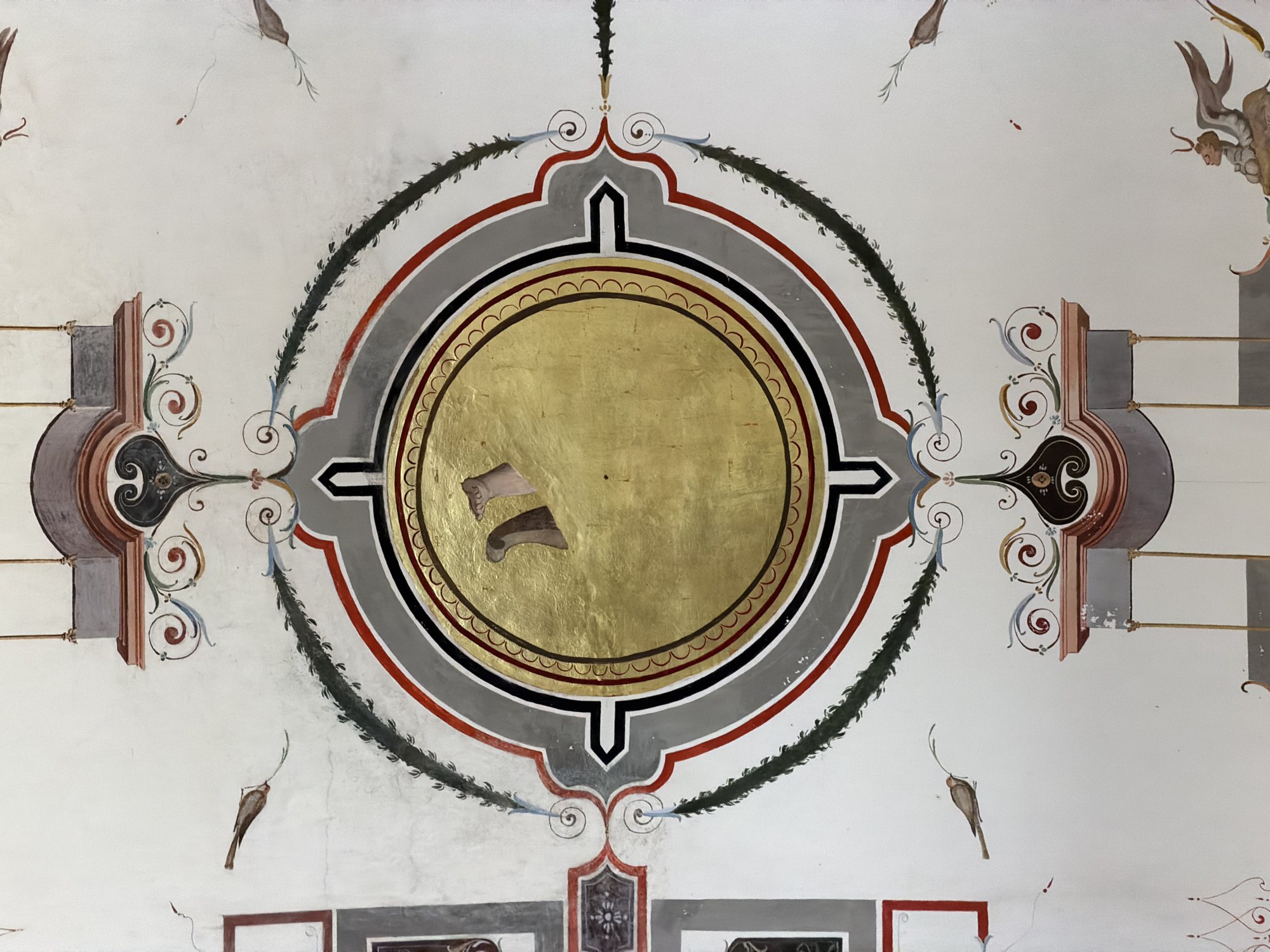 This image shows a fragment of a frescoed ceiling from San Lorenzo de El Escorial, Spain. The central focus is a large, circular motif with a golden, textured background, possibly representing a medallion or a celestial body. This central circle is surrounded by several concentric rings of painted decoration, featuring bands of black, red, and grey, with a scalloped inner edge. The entire circular design is framed by ornate architectural elements and decorative scrolls in shades of grey, brown, and red.
Flanking the central medallion are stylized, architectural elements that resemble corbels or brackets, adorned with swirling patterns and darker accents. Extending from these architectural pieces are thin, decorative vines or branches with dark green, spiky leaves. These vines arc outwards and upwards, framing the central design.
Scattered across the white background of the ceiling are small, painted figures of birds, rendered in a simple, illustrative style. There are also some scattered red dots and faint lines, possibly remnants of other decorative elements or imperfections in the fresco. At the top right of the image, a small, winged figure, possibly an angel or cherub, is partially visible, with brown skin and wings.
The fresco appears to be aged, with some areas of wear and slight discoloration, indicating its historical nature. The overall style is decorative and somewhat formal, characteristic of Renaissance or Baroque period interiors. There is no discernible text within the visible portions of the image. The setting is clearly an interior, likely a grand hall or chapel within a historic building.