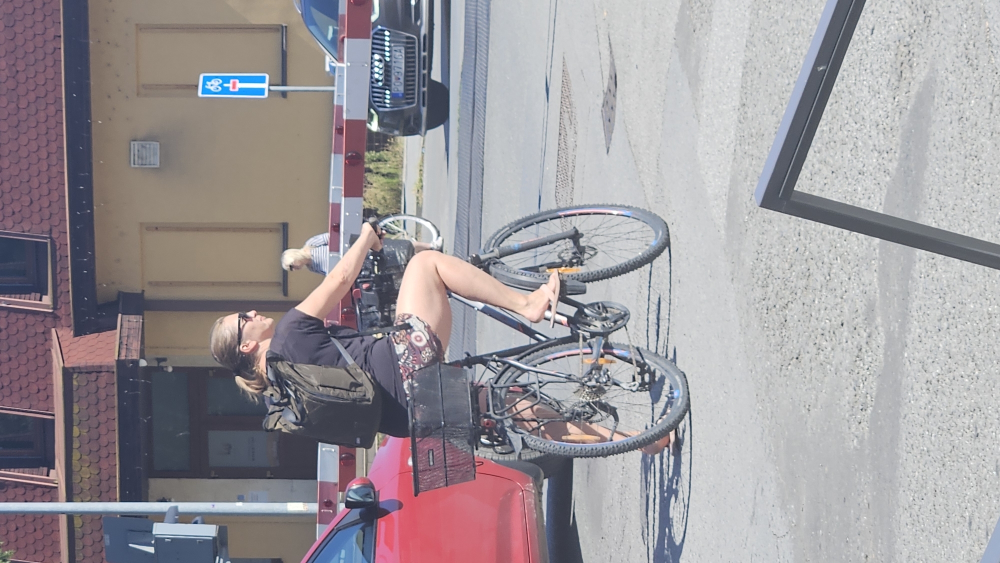 A woman wearing sunglasses, a dark t-shirt, and patterned shorts is riding a bicycle with a backpack. Her right foot is in a flip-flop, while her left foot is bare. The bicycle has a basket on the back containing some items. The woman is riding across a railway crossing with the barriers down. A red car is parked to the left of the crossing, and a dark Audi car is visible on the other side, stopped at the barrier. A woman with blonde hair is standing in the background near a building. A bicycle sign is mounted on a pole on the right side of the image. The scene appears to be in Prešov, Slovakia.