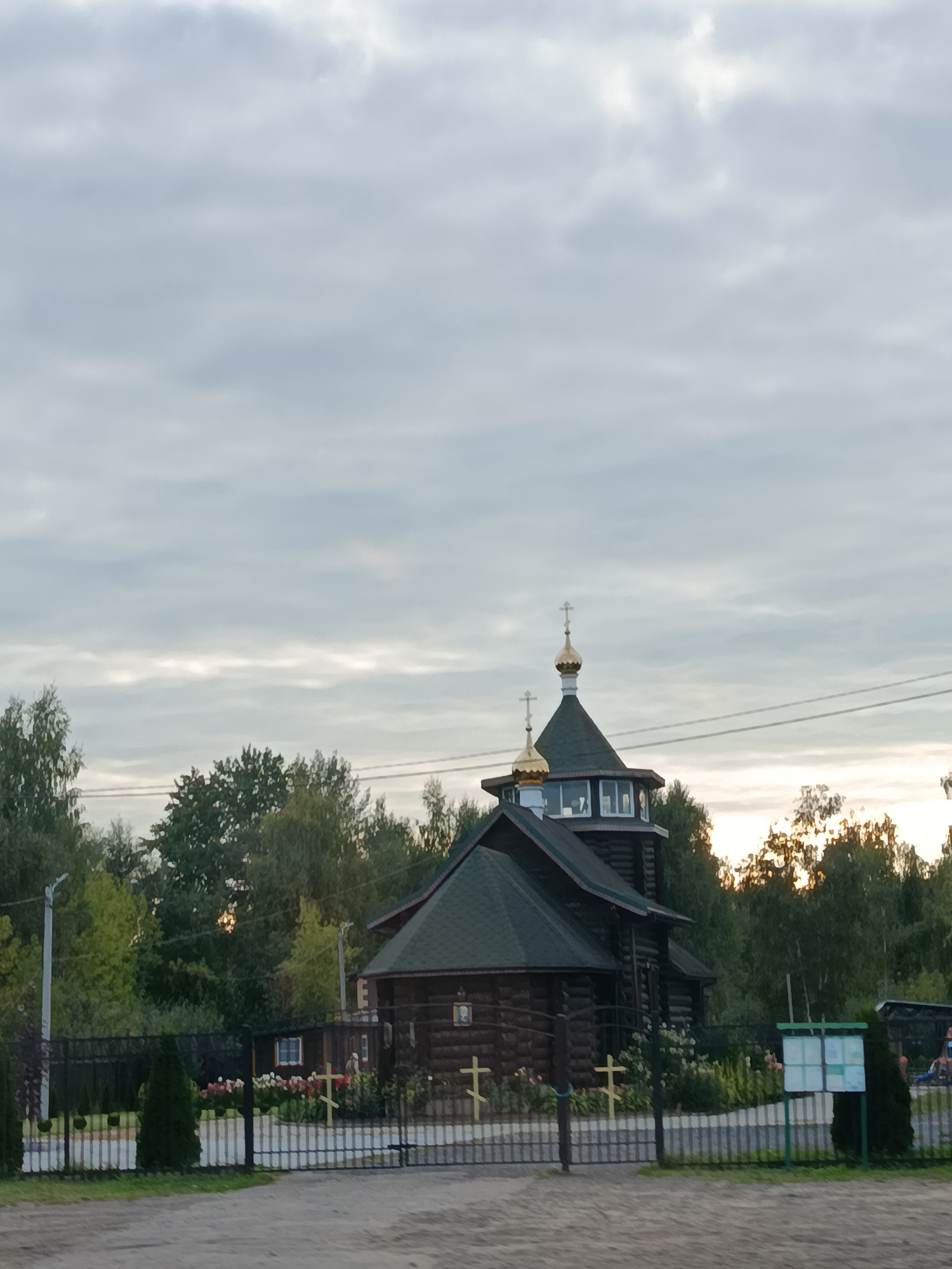 The image shows a wooden church with a green roof and golden domes topped with crosses. The church is situated amidst trees and a fence. The sky is overcast with clouds. The overall scene suggests a rural or suburban setting. The church appears to be a place of worship, possibly a Russian Orthodox church given its architectural style. There are no people visible in the image. The environment is natural, with abundant greenery, and the weather appears to be cloudy, possibly during the day or early evening. The image does not provide any specific landmarks or details that would definitively pinpoint its location within the city of Vereya, Russia, beyond it being a church in that city.