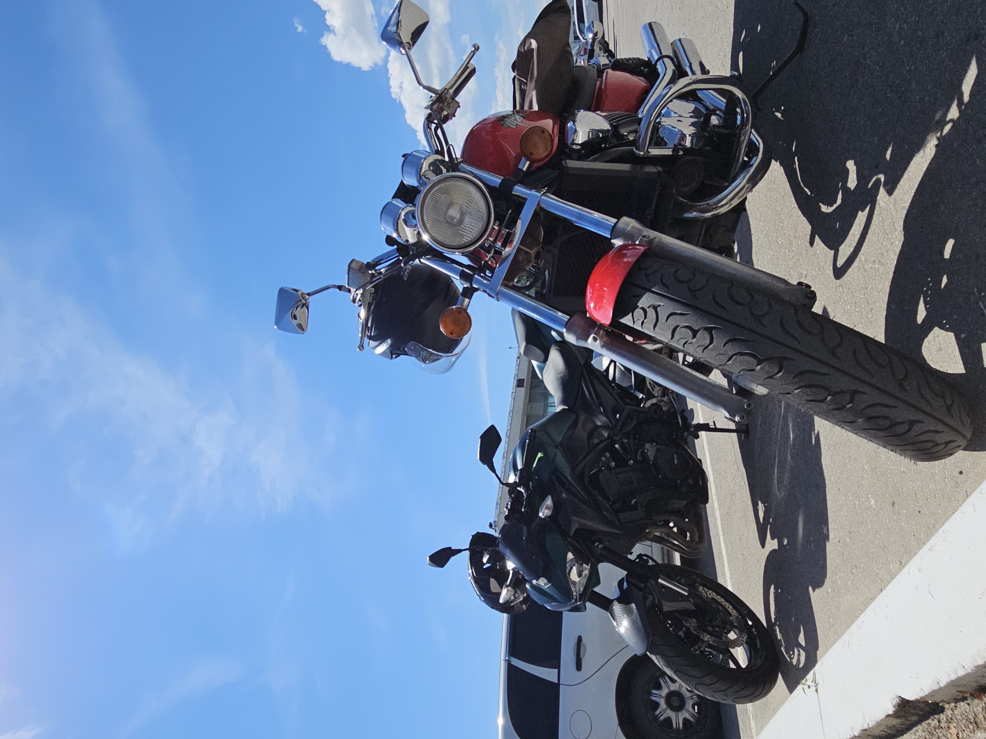 Two motorcycles, one red and one dark green, are parked side-by-side outdoors under a clear blue sky with scattered clouds. The red motorcycle is in the foreground, facing left, with its front wheel prominently displayed. The dark green motorcycle is behind it, also facing left. A white vehicle, possibly a limousine or van, is parked to the left of the green motorcycle. The scene appears to be a parking lot, with asphalt ground and a concrete curb in the lower left corner. The image captures a sunny day. The location is identified as being in Ulyanovsk, Russia.