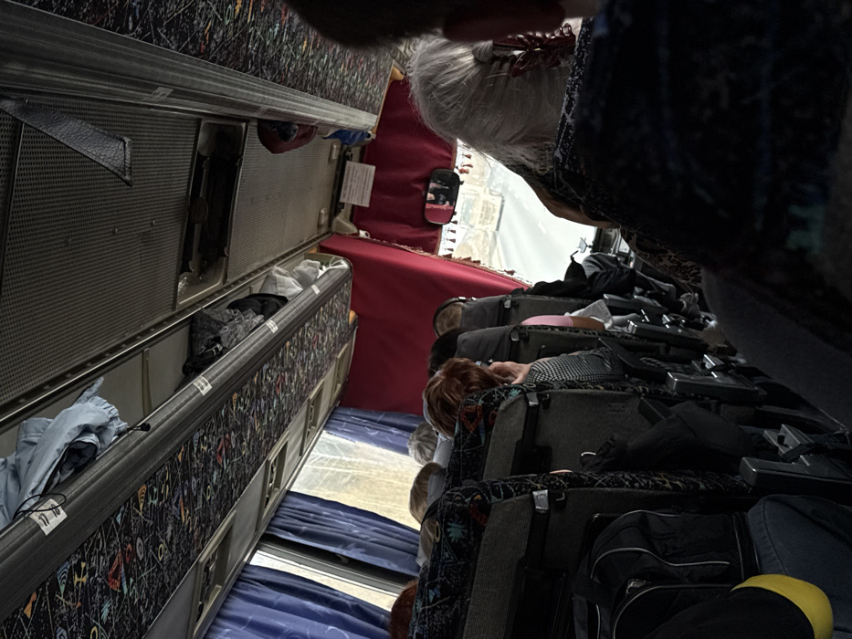 The image depicts the interior of a passenger bus in the city of Lipovka, Russia, viewed from a slightly elevated position towards the front. Multiple passengers are visible, primarily from behind, seated in rows of dark, patterned fabric seats. Visible passengers include individuals with reddish-brown, blonde or light-colored, and grey or white hair. No specific interactions between passengers are evident; they are generally oriented forward.
Overhead compartments run along both sides of the bus interior, some of which are open, revealing stored items such as light blue clothing and dark bags. The ceiling features a patterned fabric design. Blue curtains are drawn on the windows along the left side of the bus, while a view through the front windshield shows an asphalt road and a light-colored truck in the distance. Red curtains frame the front section of the bus, and a rearview mirror is visible above the driver's area. A white rectangular sign is mounted near the front ceiling, above the windshield. A black bag with a yellow reflective element is situated near the foreground on the left side of the aisle. The overall lighting suggests an overcast outdoor environment.