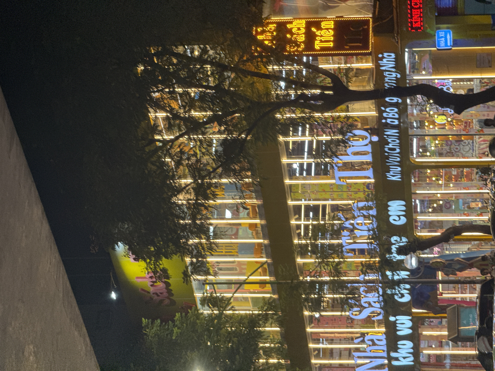 This is a nighttime photograph of a multi-story building in Hanoi, Vietnam, likely a bookstore and entertainment center. The building is illuminated with prominent neon signs. The largest sign on the upper facade reads "Sách" in pink. Below this, a large white neon sign spells out "Nhà Sách Tiên Thọ." Beneath that, another white neon sign says "Khu Vui Choi Nà Bo g ong Nhà," translating to "Children's Play Area." The building's facade is mostly glass, allowing glimpses of the interior, which is also lit and appears to contain displays and seating areas. Trees with sparse foliage are in the foreground, partially obscuring the building. The overall impression is of a brightly lit commercial establishment at night in an urban environment.