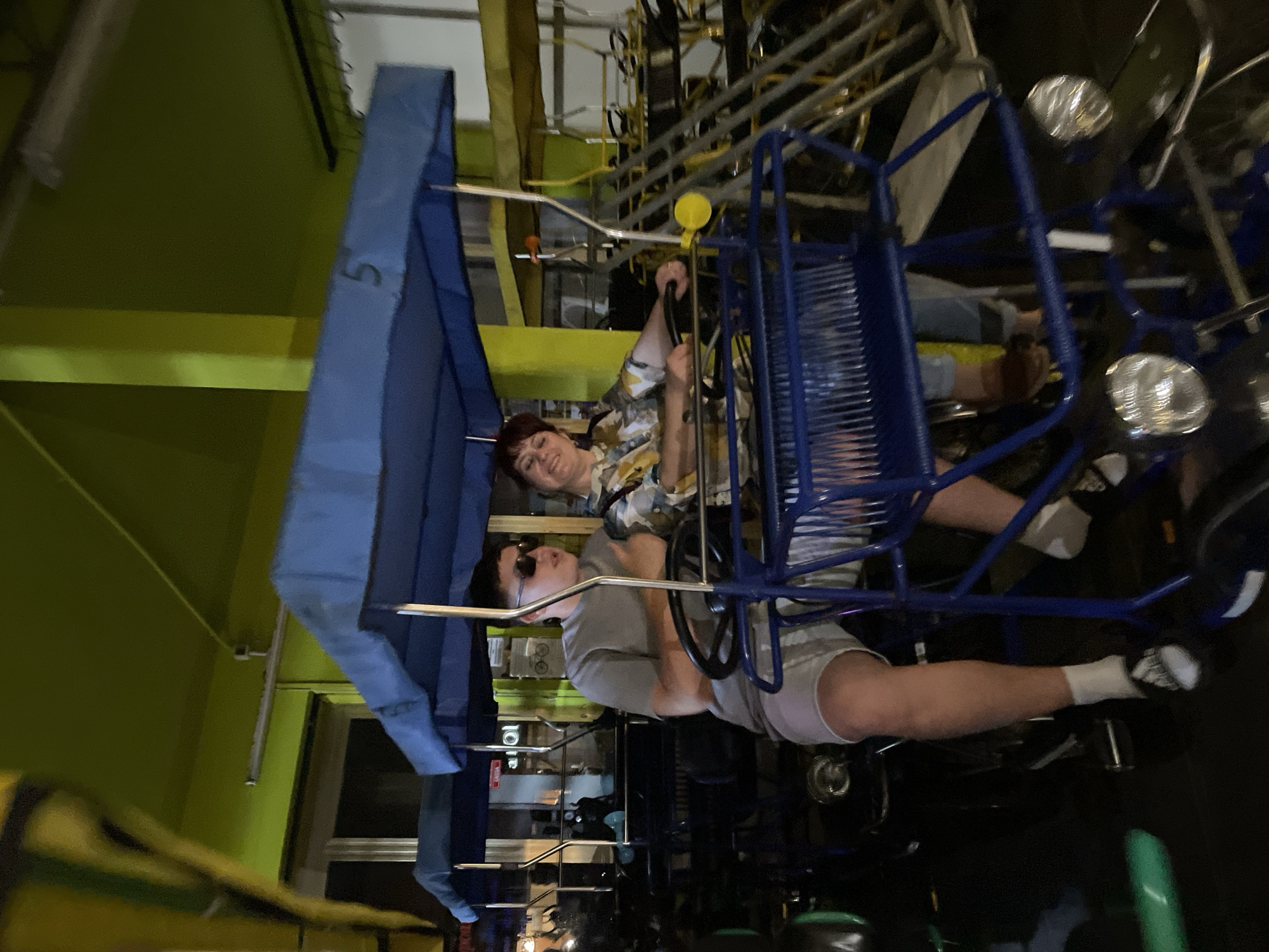 Two people are seated in a blue, quadricycle-like vehicle. The person in the front wears sunglasses and a light grey t-shirt with grey shorts, white socks and black shoes. The person in the back wears a floral shirt. The setting appears to be indoors, perhaps an amusement park or a tourist attraction. The background includes a green wall on the left and metal structures. Number "5" appears to be written on the top left. The precise location within Jesolo, Italy, is not explicitly indicated within the image.