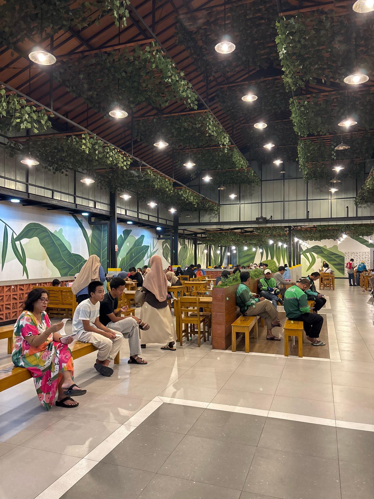 The image depicts an indoor dining area, possibly a food court or restaurant, in Bekasi, Indonesia. The space features a high ceiling adorned with artificial green vines and hanging lamps. The walls are decorated with a mural of large green leaves. Numerous people are seated at tables and on benches, some appearing to be waiting for food or companions. The flooring is tiled with light and dark grey squares, separated by white lines. Several individuals are wearing green jackets with the "Grab" logo, indicating they might be delivery drivers. The lighting suggests it is evening or indoors with artificial illumination.