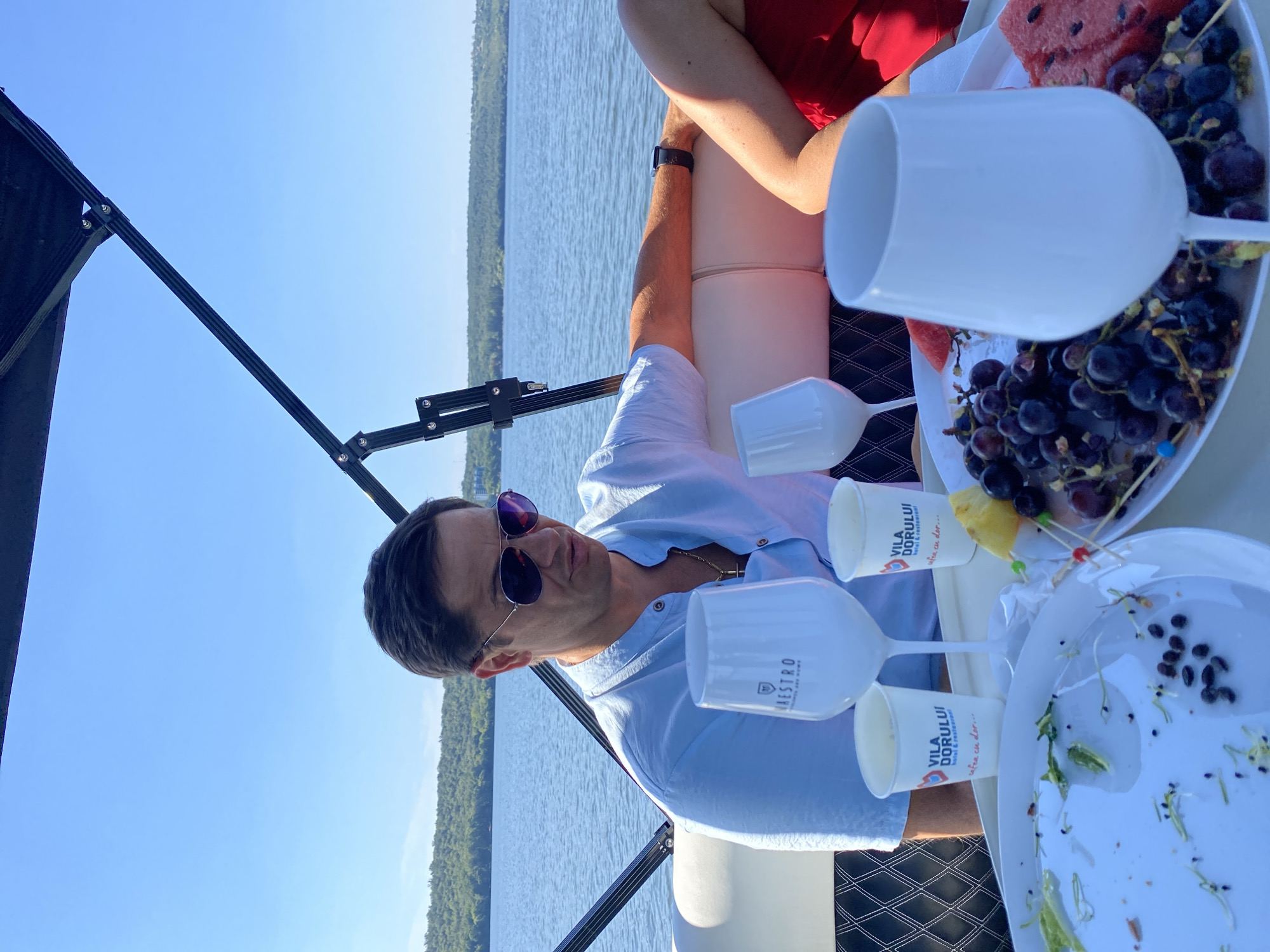 A man in a light blue collared shirt and sunglasses sits on a boat, looking down. He wears a gold chain and a watch on his left wrist. In front of him is a table with white plates of fruit, grapes, and watermelon, as well as white cups and wine glasses. The boat is on a body of water, with a tree-lined shore visible in the distance under a clear blue sky. This scene is located in the city of Korzhevo, Moldova.