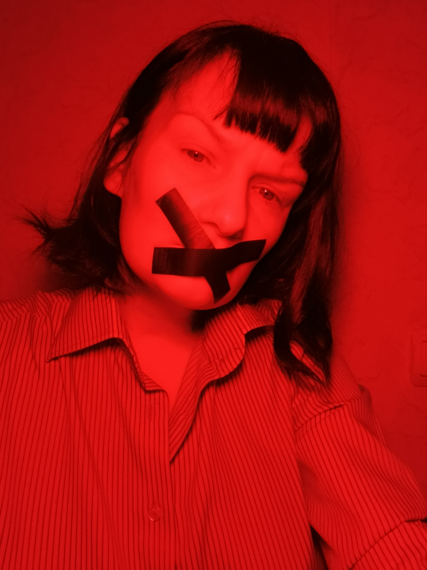 A woman is shown in a close-up shot. Black tape covers her mouth in an "X" shape. She is wearing a striped button-down shirt. The lighting is red, giving the image a monochromatic appearance. The scene's location is not discernible from the image.