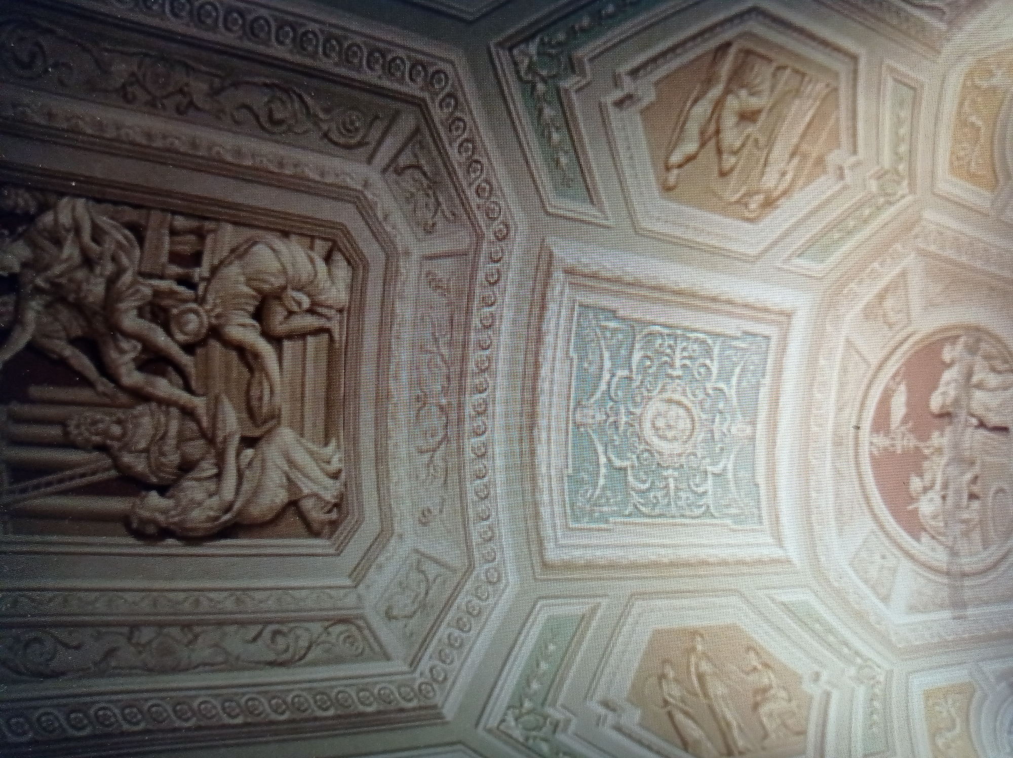 The image shows a detailed, ornate frescoed ceiling, likely in a historical building. The ceiling features intricate patterns, geometric designs, and relief sculptures depicting human figures in various poses. The style appears to be classical or Renaissance. There are no people visible in the image, nor are there any indications of the city of Mukachevo, Ukraine. The focus is solely on the architectural and artistic elements of the ceiling.