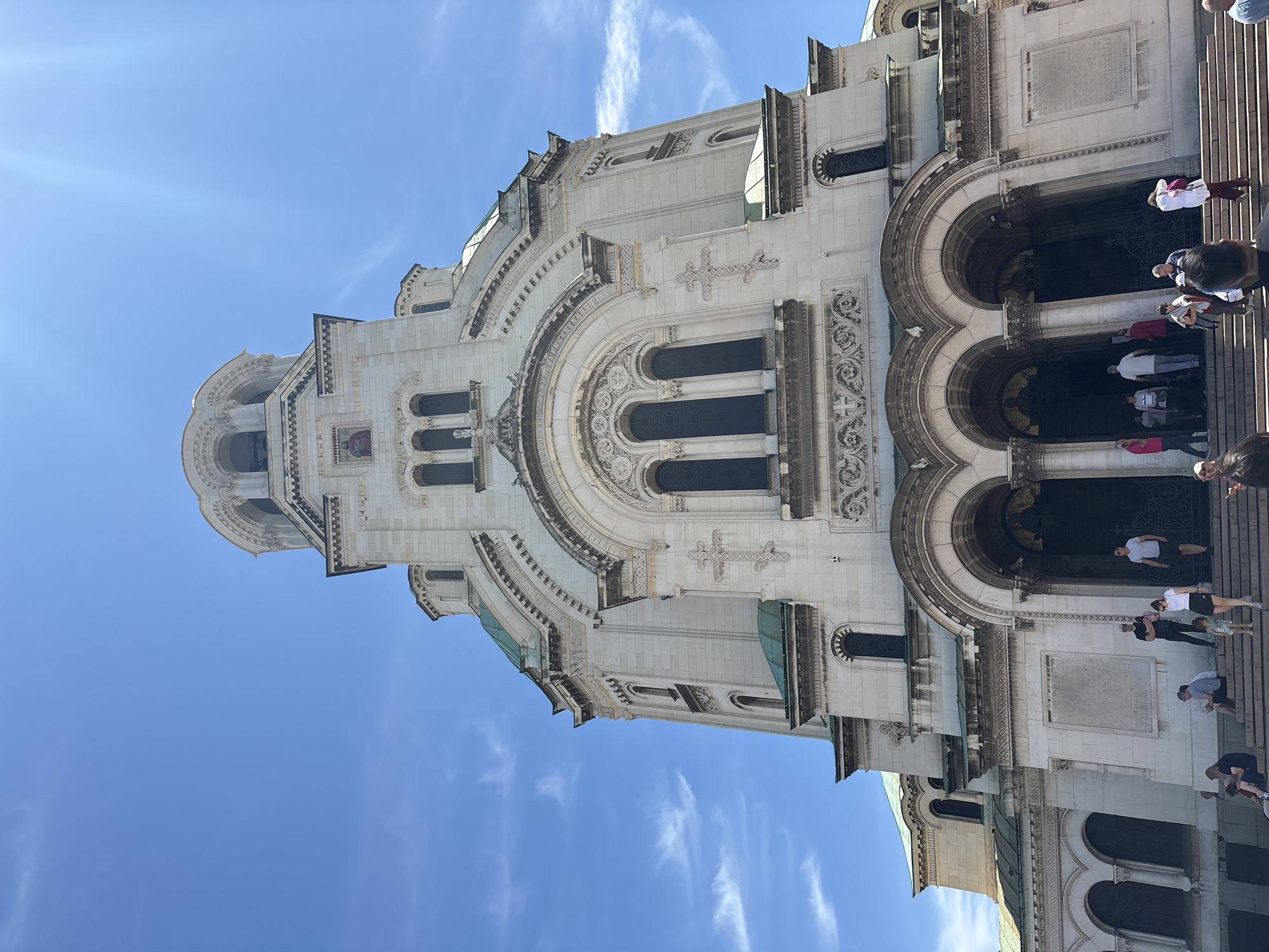 The image shows the Alexander Nevsky Cathedral in Sofia, Bulgaria. The cathedral is constructed of white stone and features multiple arches, windows, and decorative elements such as crosses and intricate carvings. The sky is blue with thin, scattered clouds. A group of people can be seen on the steps leading to the cathedral's entrance. Some are entering the building, and others are standing on the steps.