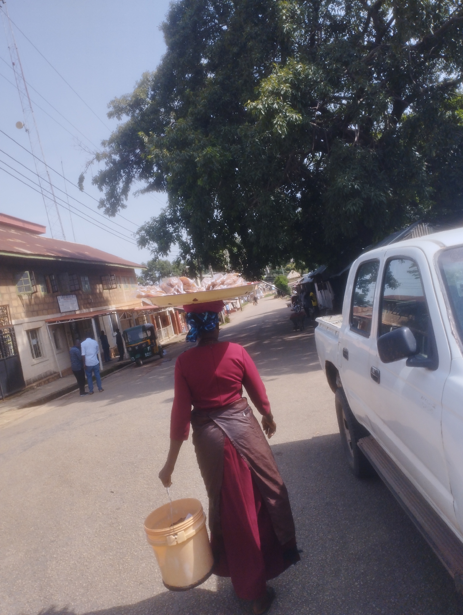 A woman is walking down a paved road, carrying a large tray of food items on her head. She is wearing a maroon top and a brown skirt, and a blue headscarf. She is also carrying a yellow bucket in her right hand. In the background, there is a white pickup truck, a building with a red roof, a large tree, and power lines. There are a few other people walking in the distance. The scene appears to be set in a town or city in Jos, Nigeria, during the daytime. The weather appears to be sunny and clear. There is no visible text in the image.
