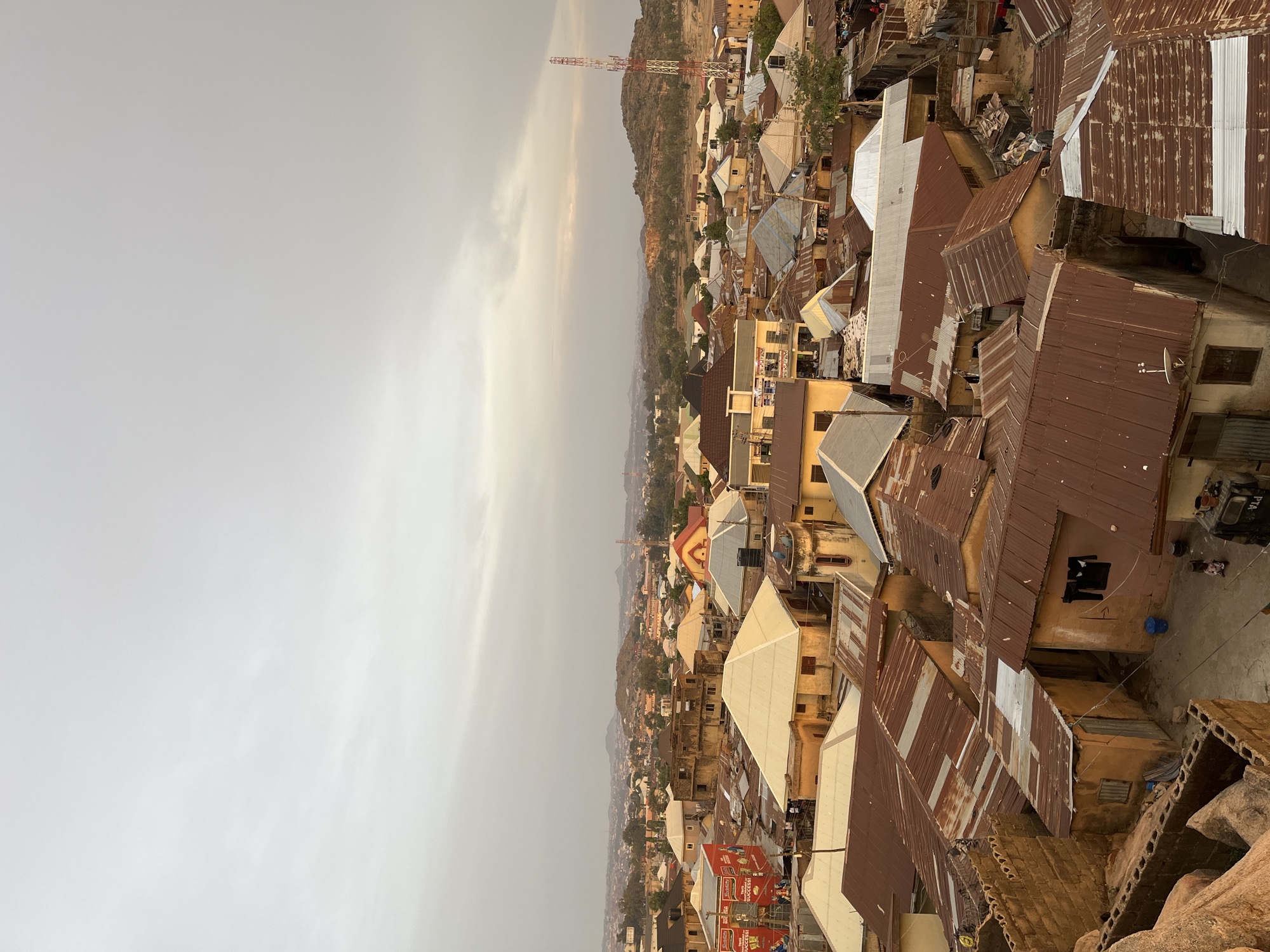 The image depicts a high-angle view of a densely populated urban area in Jos, Nigeria. The foreground shows rooftops, predominantly made of corrugated metal, with variations in color ranging from brown to silver. Some buildings have visible walls and windows. In the middle ground, there are more buildings and structures of varying heights and colors. A communications tower is visible against a hilly horizon. The background shows the sky, which is overcast and appears hazy. People are faintly seen, and some objects like satellite dishes are also visible. The overall environment suggests an urban residential area.