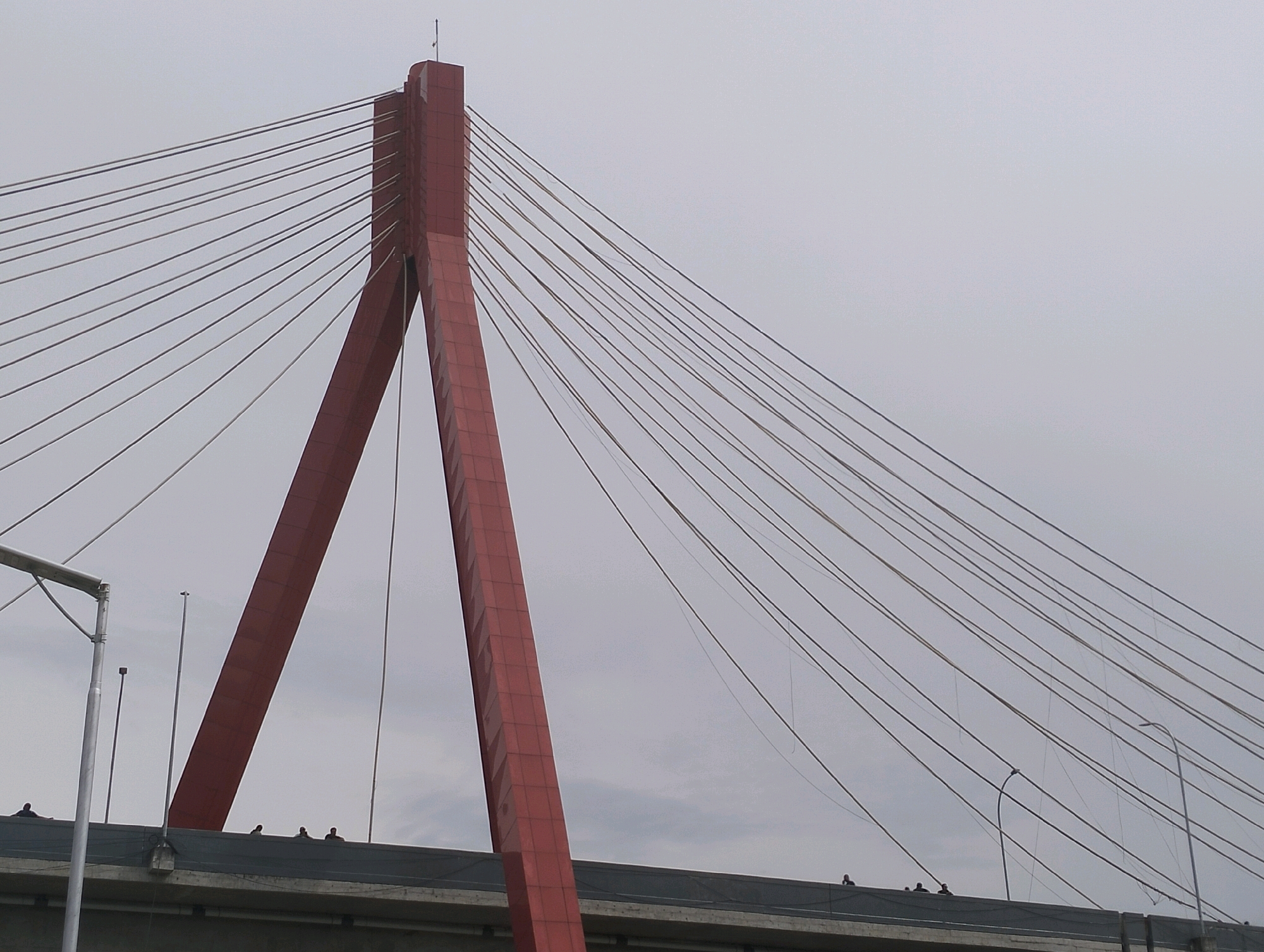 The image shows a close-up view of a red cable-stayed bridge tower against a cloudy sky in Siogung-Ogung, Indonesia. The tower is a tall, angular structure with multiple cables radiating outwards to support the bridge deck. Several thin vertical poles, likely streetlights or antenna mounts, are visible on the bridge deck. Silhouetted figures of people can be seen standing on the bridge deck near the base of the tower and further along its length. The environment appears to be overcast, with a diffused light.