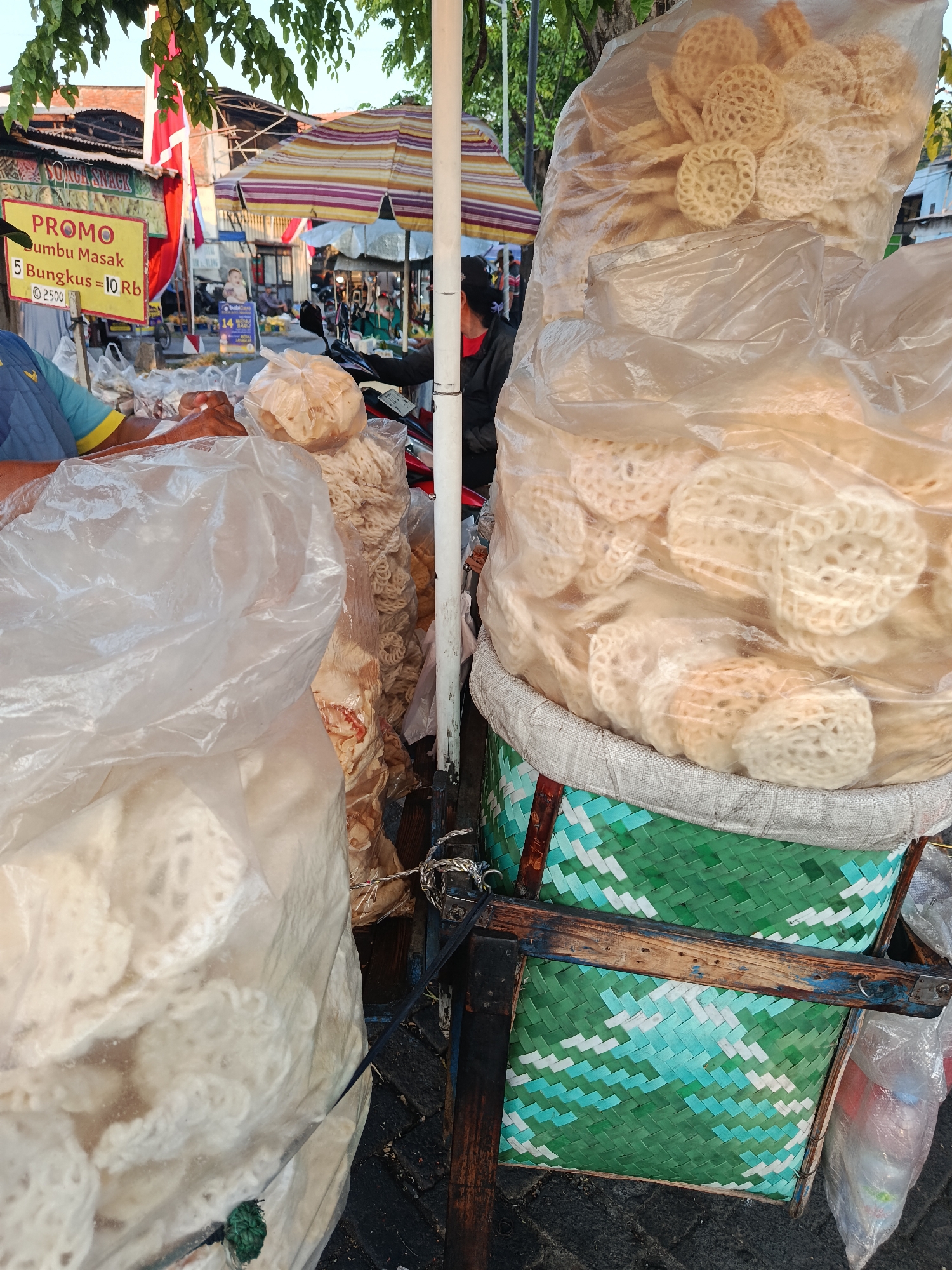 A street vendor in Keboansikep, Indonesia, is captured in a close-up shot, showcasing their wares. The foreground is dominated by large, transparent plastic bags filled with a variety of crackers, some circular and intricately patterned, others long and thin. These are stacked and secured with rope. Behind the crackers, a woven basket with a green and white diamond pattern is visible, further supporting the stacked bags of crackers. A wooden frame supports the basket and bags, suggesting a cart or stall. A sign with the text "PROMO" and Indonesian currency (Rp) indicates a sale on "Bumbu Masak." In the background, glimpses of other market stalls, umbrellas, and people are visible, along with trees and buildings, suggesting a bustling outdoor market environment. The lighting indicates it's likely daytime, possibly late afternoon due to the warm, soft light.