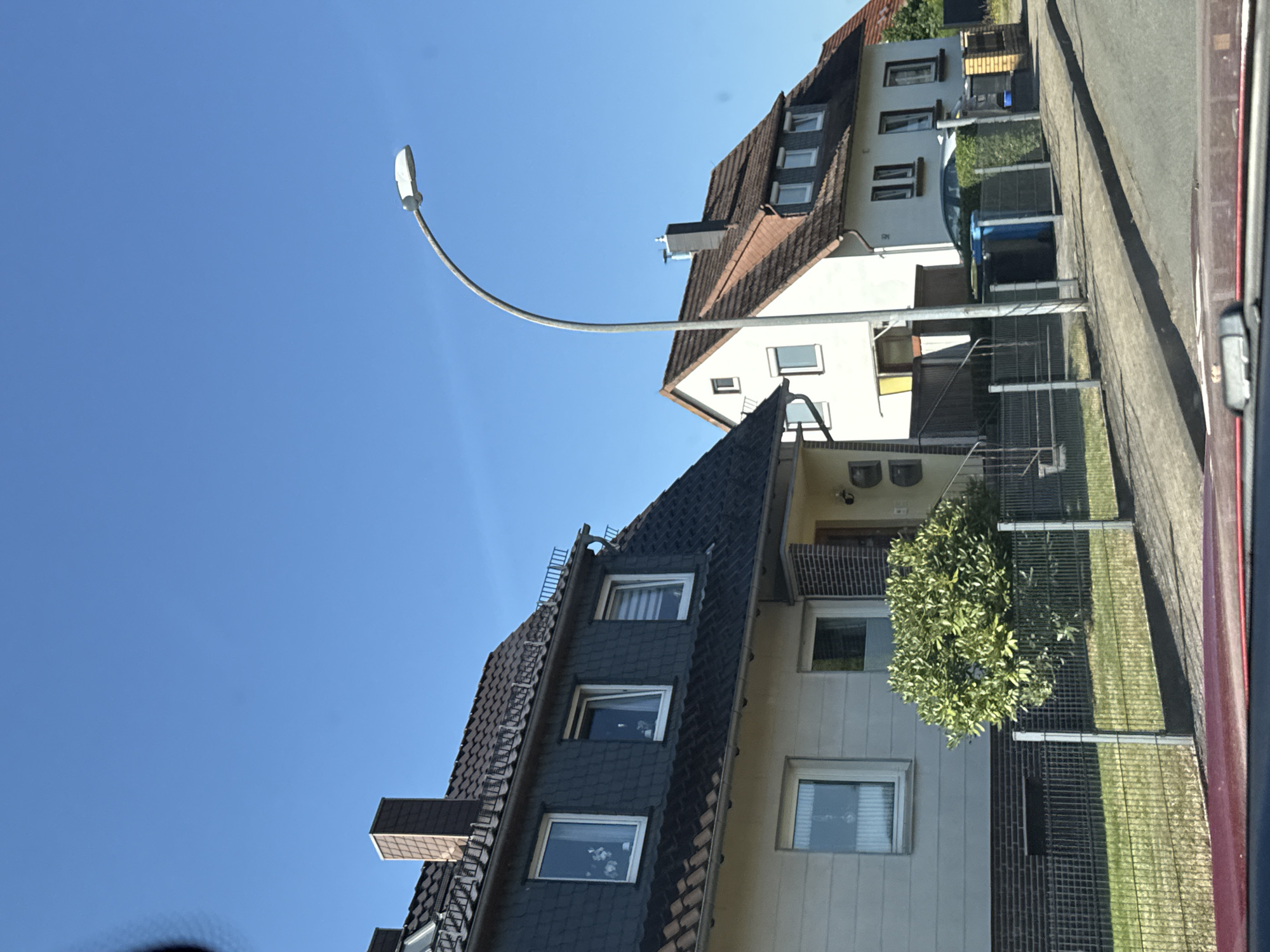 The image depicts a residential street in Bad Sachsa, Germany, under a clear blue sky. Two houses are visible, with the house on the left featuring a grey slate facade on its upper level and a cream-colored lower level. This house has multiple windows, some with white frames and others with darker frames, and a dark-colored tiled roof with a prominent chimney. A small, bushy green tree is situated in front of this house, behind a metal fence.
The house on the right is white with a red-tiled roof and several dormer windows. A dark-colored car is parked in front of this house, partially obscured by a green hedge and a metal fence. Two trash bins, one blue and one black, are visible behind the fence in front of the house. A modern street lamp with a curved arm arches over the scene. The foreground shows the edge of a road and what appears to be the interior of a vehicle, suggesting the photo was taken from a car. The overall impression is a quiet suburban street scene on a sunny day.