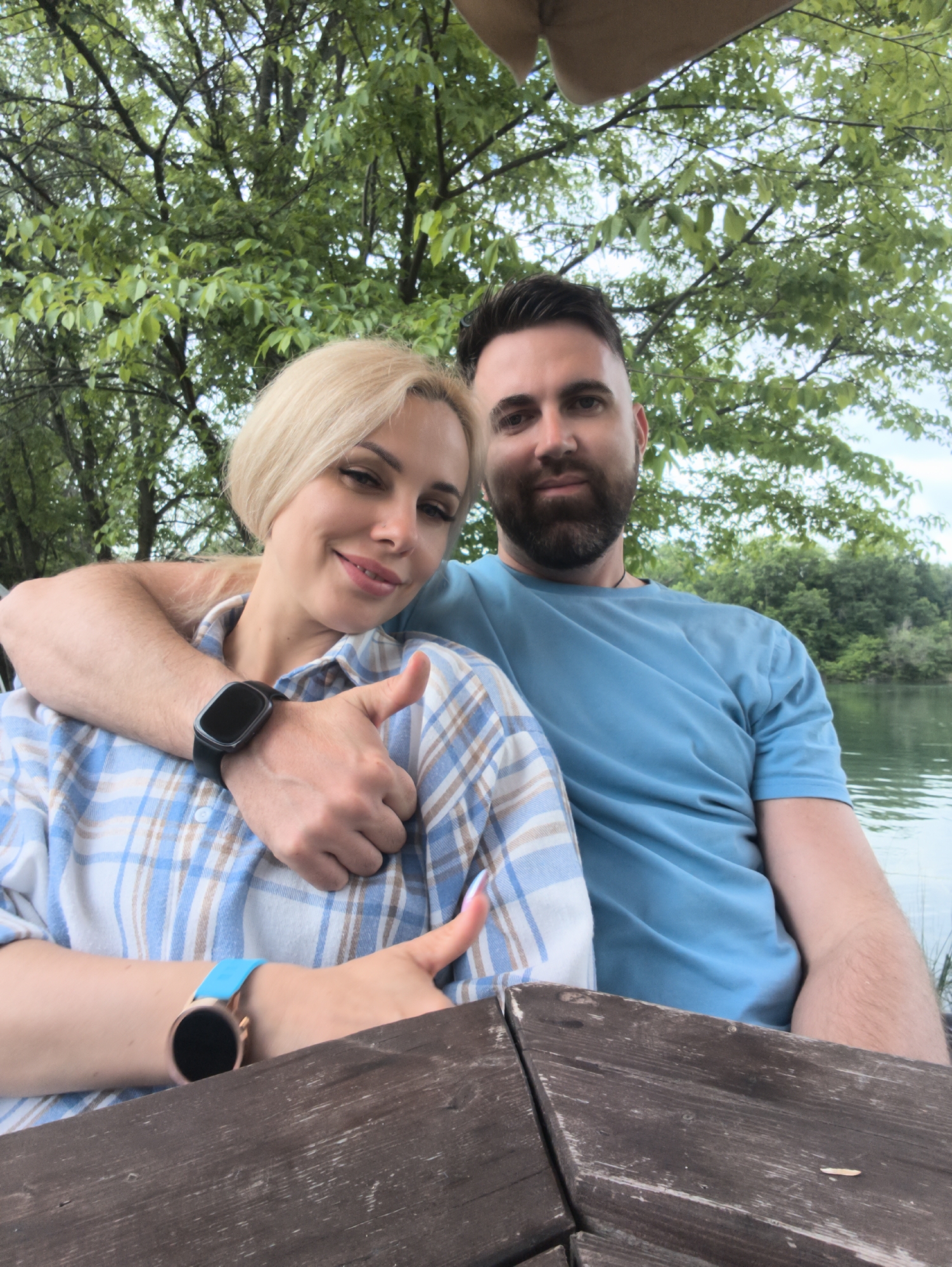 A blonde woman in a blue plaid shirt sits at a wooden picnic table with a bearded man wearing a blue t-shirt. The man has his arm around the woman. Both are giving a thumbs-up gesture. The woman is wearing a smartwatch with a blue wristband, and the man is wearing a smartwatch with a black wristband. Behind them, there are trees and a body of water. The location appears to be Терновский округ, Ukraine.