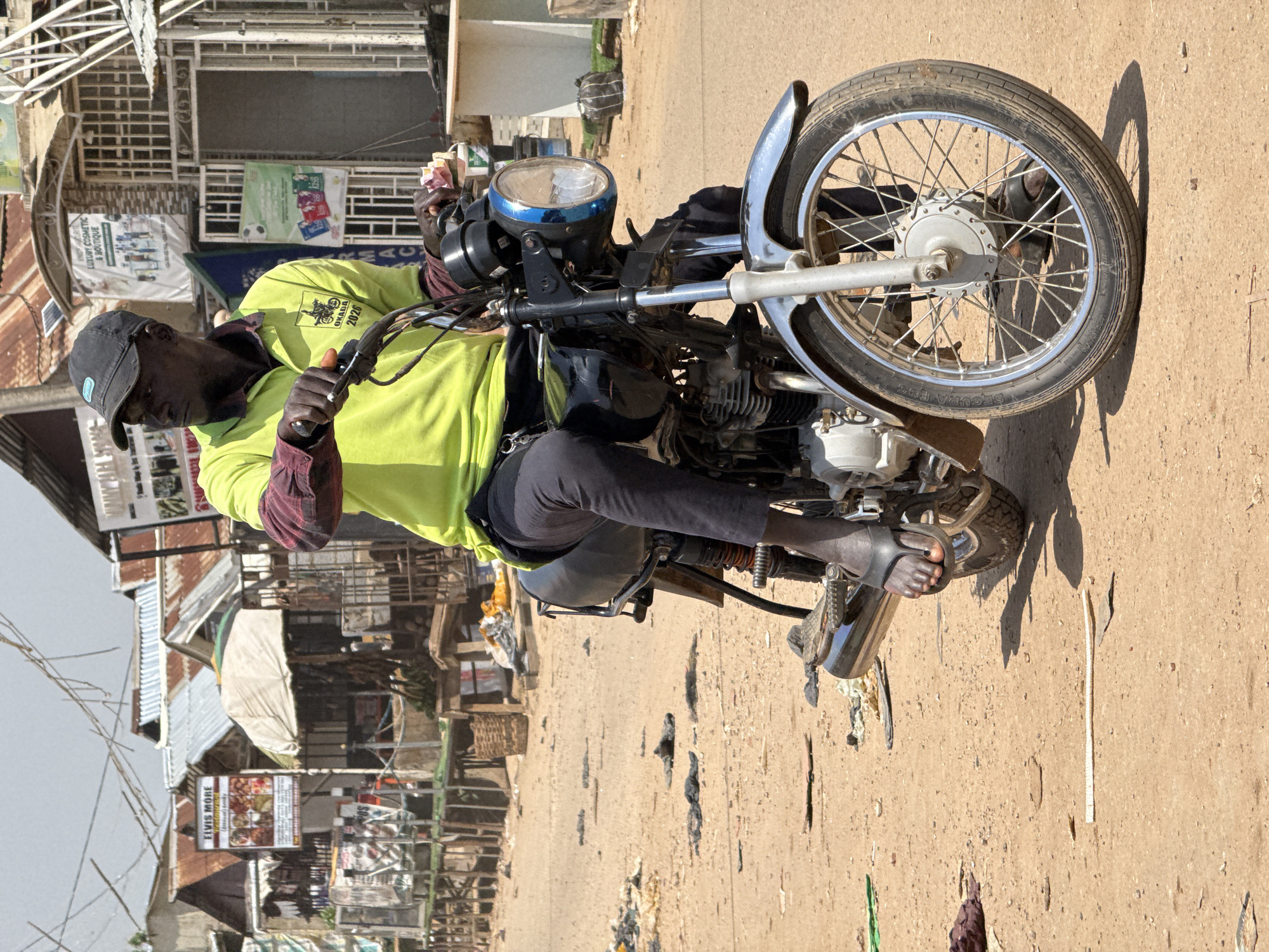 A dark-skinned man wearing a lime green polo shirt, a maroon long-sleeved shirt underneath, dark pants, a black baseball cap, and flip-flops is seated on a black motorcycle. He is looking to his left. The motorcycle has a chrome fender and a blue and chrome headlight. The man is holding the handlebars with his right hand and appears to be holding some pink paper money in his left hand. The motorcycle is parked on a dirt road with scattered debris. In the background, there are buildings with corrugated metal roofs, some of which appear to be shops or market stalls. One sign visible says "ELVIS MORE RESTAURANT". The scene appears to be an outdoor market or commercial area in Abuja, Nigeria.