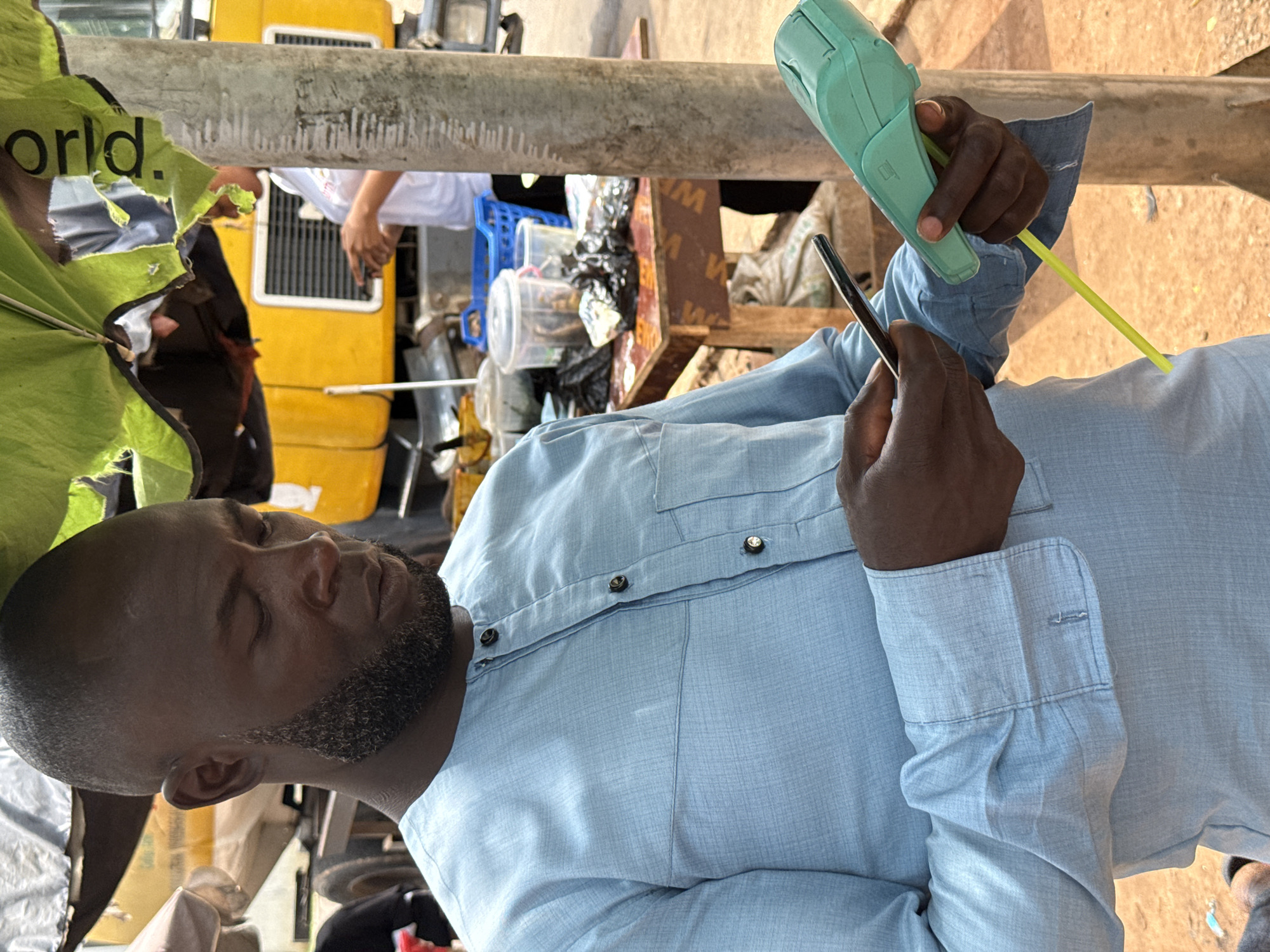 A dark-skinned man with a shaved head and a goatee is standing outdoors in what appears to be a market or street vending area in Abuja, Nigeria. He is wearing a light blue, long-sleeved tunic with dark buttons. He is holding a smartphone in his left hand and a teal-colored card reader with a yellow cable attached in his right hand, and he is looking down at the phone. In the background, a yellow vehicle, possibly a truck, is visible. There are also various containers and bags on a wooden table to the man's left, suggesting a commercial transaction. The man's skin is dark brown, and he has a prominent beard. The environment appears to be somewhat crowded and dusty, with some overhead covering made of green and white fabric.