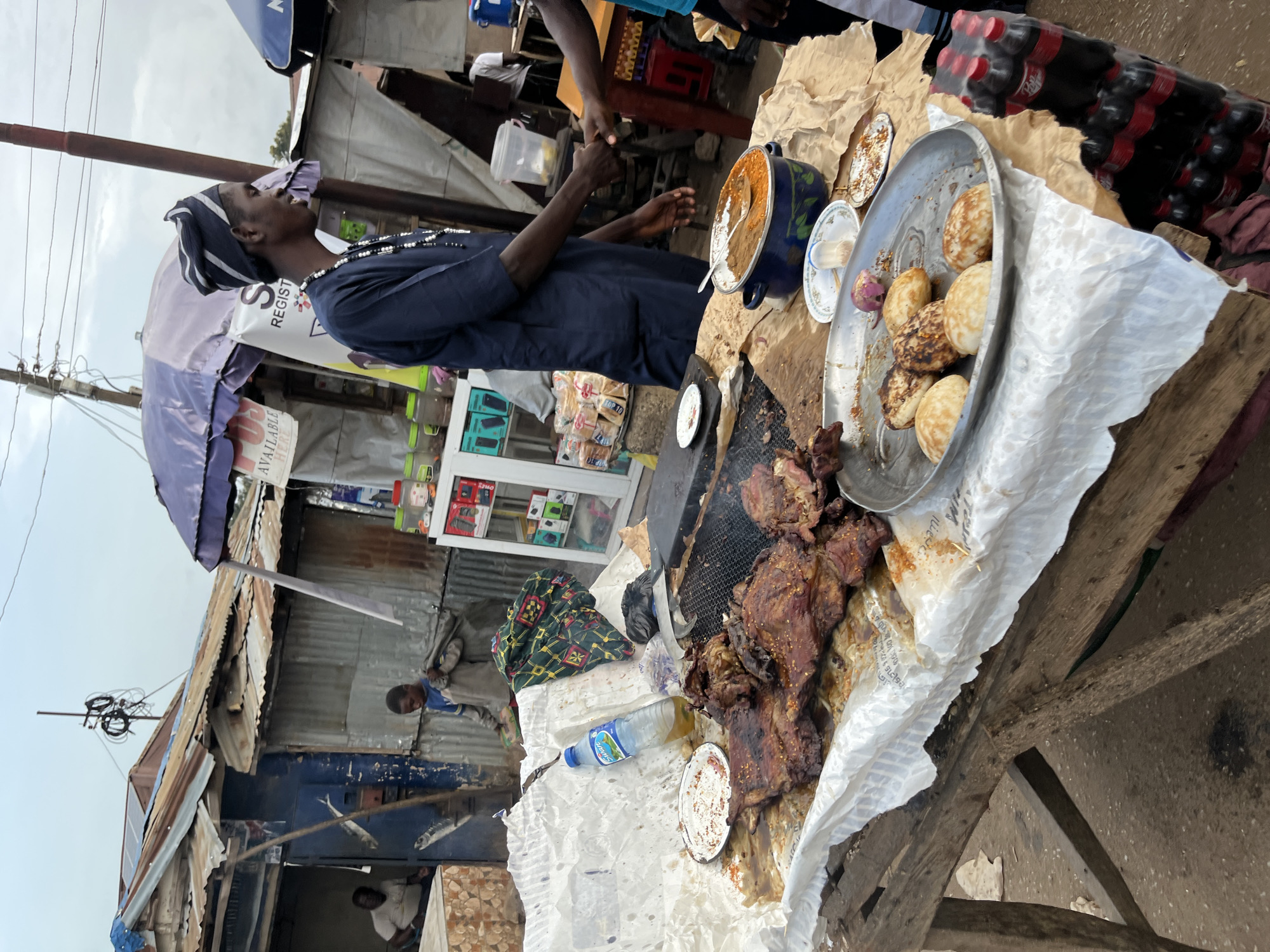 This is an outdoor street food scene in Bukuru, Nigeria. A vendor, dressed in a dark blue traditional outfit and headwrap, is seen interacting with a customer, with their hands clasped in a handshake. They are positioned in front of a makeshift stall laden with food.
In the foreground, a wooden table is covered with white paper and displays a large portion of grilled meat, appearing to be ribs or a similar cut, seasoned and cooked to a dark brown. Next to the meat is a metal platter holding several round, puffed pastries, possibly akara or puff-puff. A blue pot filled with a reddish-brown stew or sauce, with a spoon resting in it, sits on a brown paper covering the table. A bottle of water is also visible on the table.
Behind the food stall, there are various structures constructed from corrugated metal and wood, characteristic of informal markets. A young boy is walking in the background, carrying a sack over his shoulder. Further back, a man is seated inside a stall. The environment appears to be a bustling market setting, with overhead power lines and umbrellas providing shade over some of the stalls. The overall impression is that of a lively street market where food is being prepared and sold.