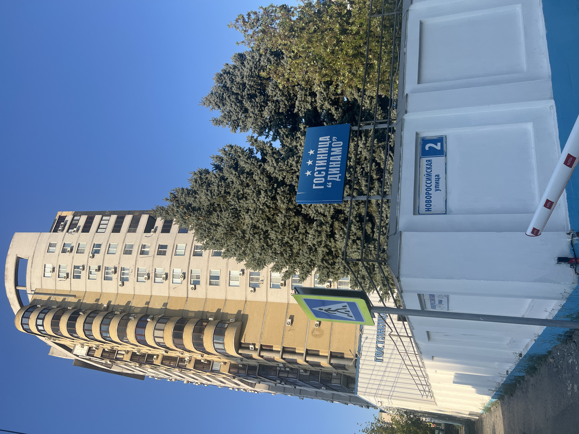 The image depicts a street scene in Volgograd, Russia, featuring a tall, modern apartment building with a curved facade, alternating between beige and cream-colored brickwork, and multiple balconies. In the foreground, a white wall with blue accents serves as a boundary. A blue sign with white Cyrillic text and three stars reads "ГОСТИНИЦА "ДИНАМО"" ("Hotel "Dynamo""). Adjacent to this, a street sign indicates "НОВОРОССИЙСКАЯ улица 2" ("Novorossiyskaya Street 2"). A pedestrian crossing sign with a blue square and a white silhouette of a person is also visible. Several large evergreen trees are positioned behind the wall, partially obscuring the lower portion of the apartment building. A paved walkway runs along the left side of the image. The sky is clear and blue. There are no people or animals visible in the scene.