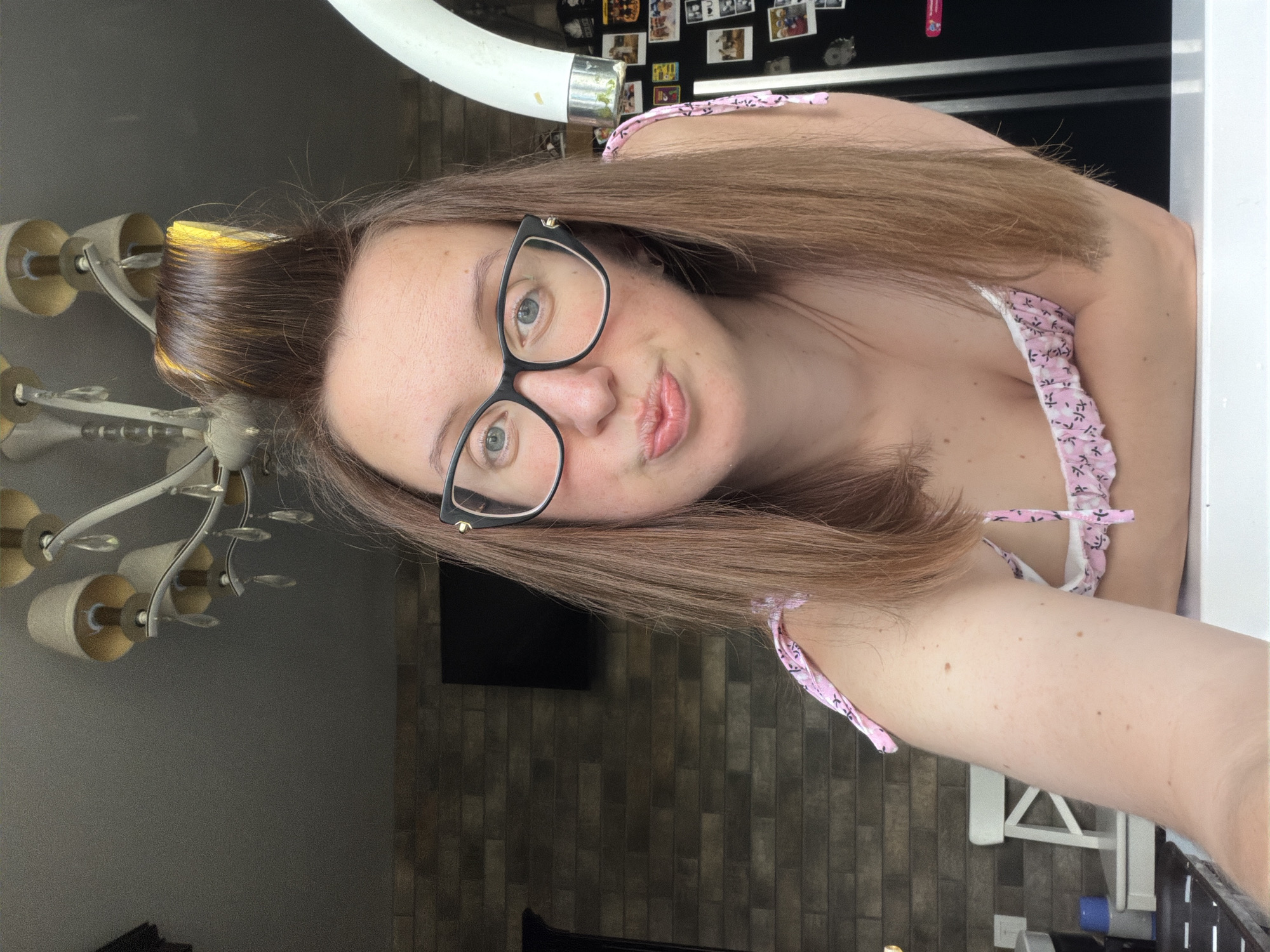 The media file is a vertical, medium shot, selfie taken indoors. The subject is a young, fair-skinned woman with medium-length, light brown hair. She is wearing black-rimmed glasses and has a light pink top with a floral pattern. Her lips are slightly puckered, and she is looking directly at the camera. The background includes a chandelier, a dark tiled wall, and a black flat-screen television. The setting appears to be a home or room. The lighting suggests it is daytime. There is no visible text in the media file. The woman's facial expression could be interpreted as playful or flirty. The overall mood is casual and personal. The user has provided a location context of Bila Tserkva, Ukraine, which is not directly discernible from the image itself but can be assumed as the location where the media file was captured.