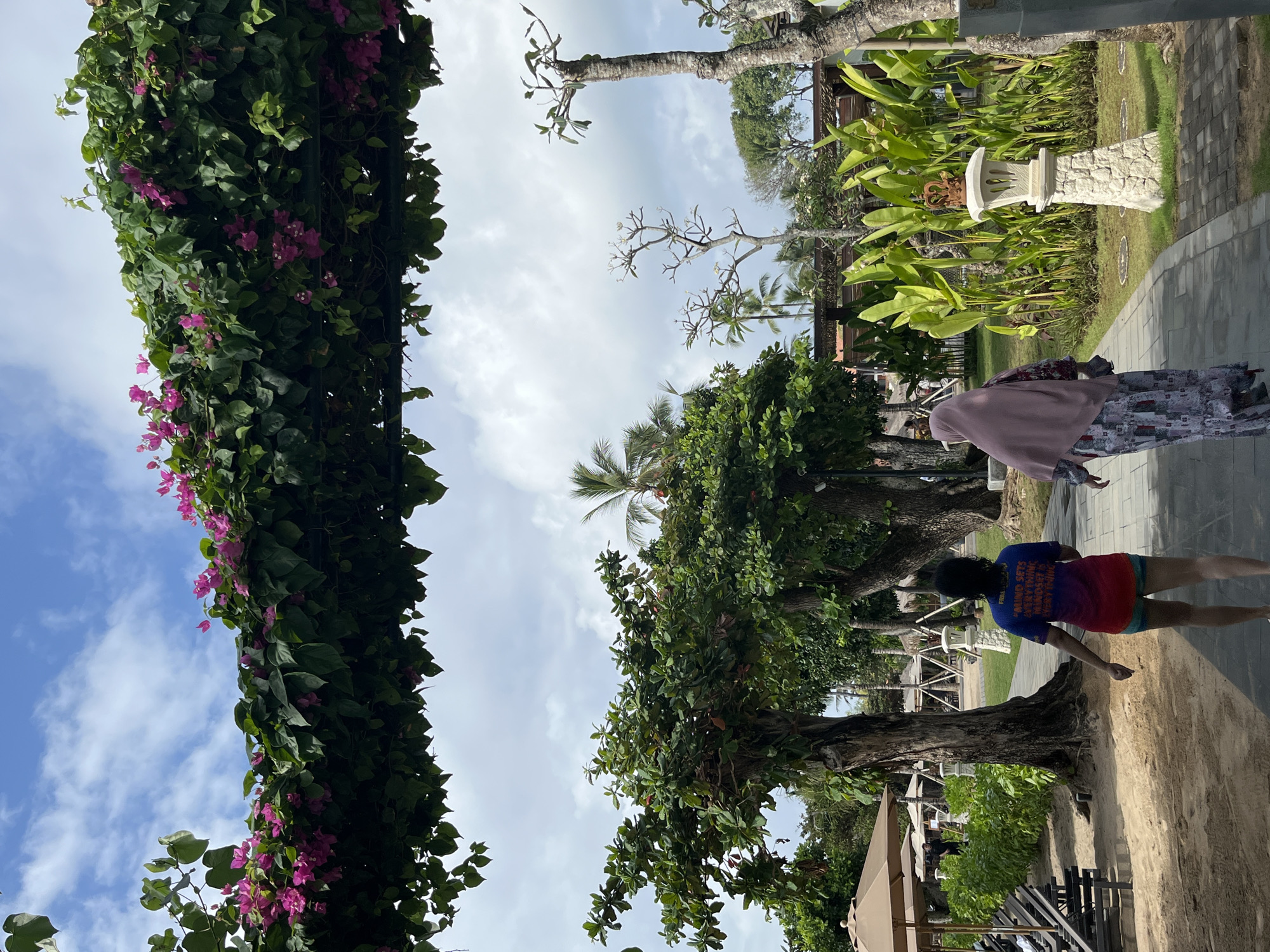In Kuta Selatan, Indonesia, a path of grey paving stones winds through a lush garden. Two women are walking away from the camera on the path. The woman on the left is wearing a t-shirt with a colorful gradient and shorts. The woman on the right is wearing a pink headscarf and a long, patterned dress.
To the left of the path, beach chairs and umbrellas are set up on a sandy area. To the right, a stone lamp post topped with a crown-like ornament stands amidst vibrant green foliage. Overhead, a canopy of green leaves and pink bougainvillea flowers arches over the scene, framed by a bright blue sky with scattered white clouds. Trees with thick trunks are interspersed throughout the garden, and palm trees are visible in the background.