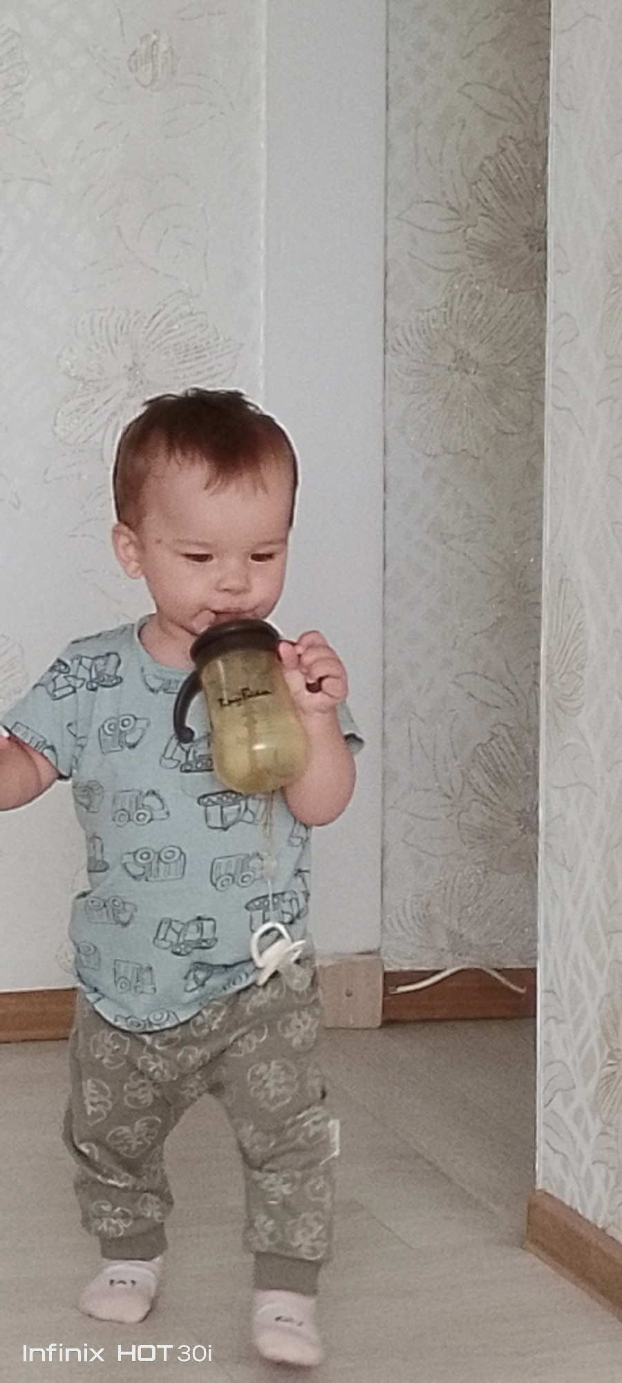 The image shows a toddler holding a sippy cup. The toddler is wearing a light blue t-shirt with a pattern of trucks and cars, and grey pants with a floral pattern. The toddler is also wearing white socks. A pacifier is attached to the t-shirt by a string. The background appears to be an indoor setting with patterned wallpaper. The location is stated to be Kedrovy, Russia.